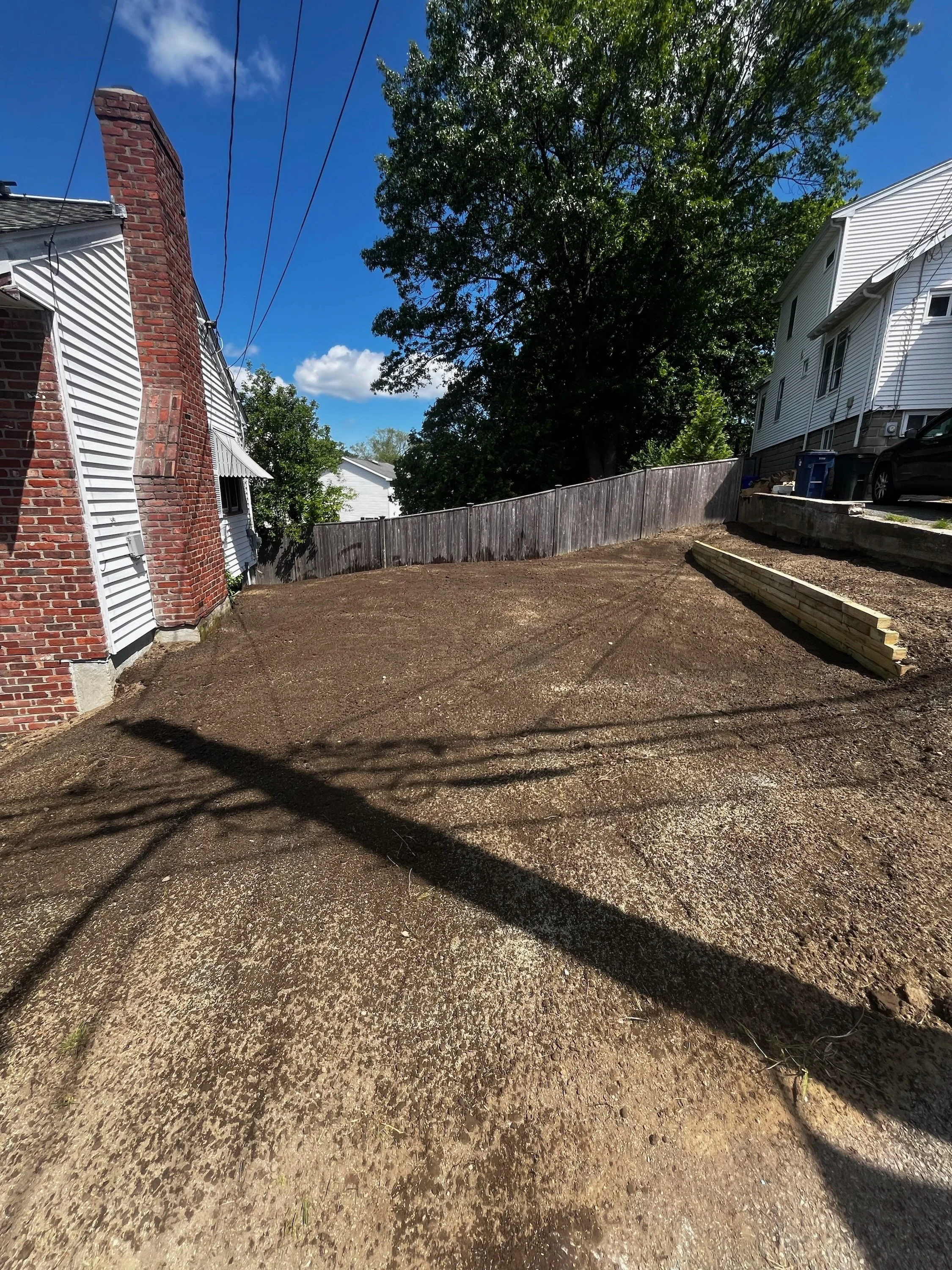 A backyard with a freshly graded dirt surface, a wooden retaining wall on the right, a tall tree in the background, neighboring houses, and a clear blue sky.