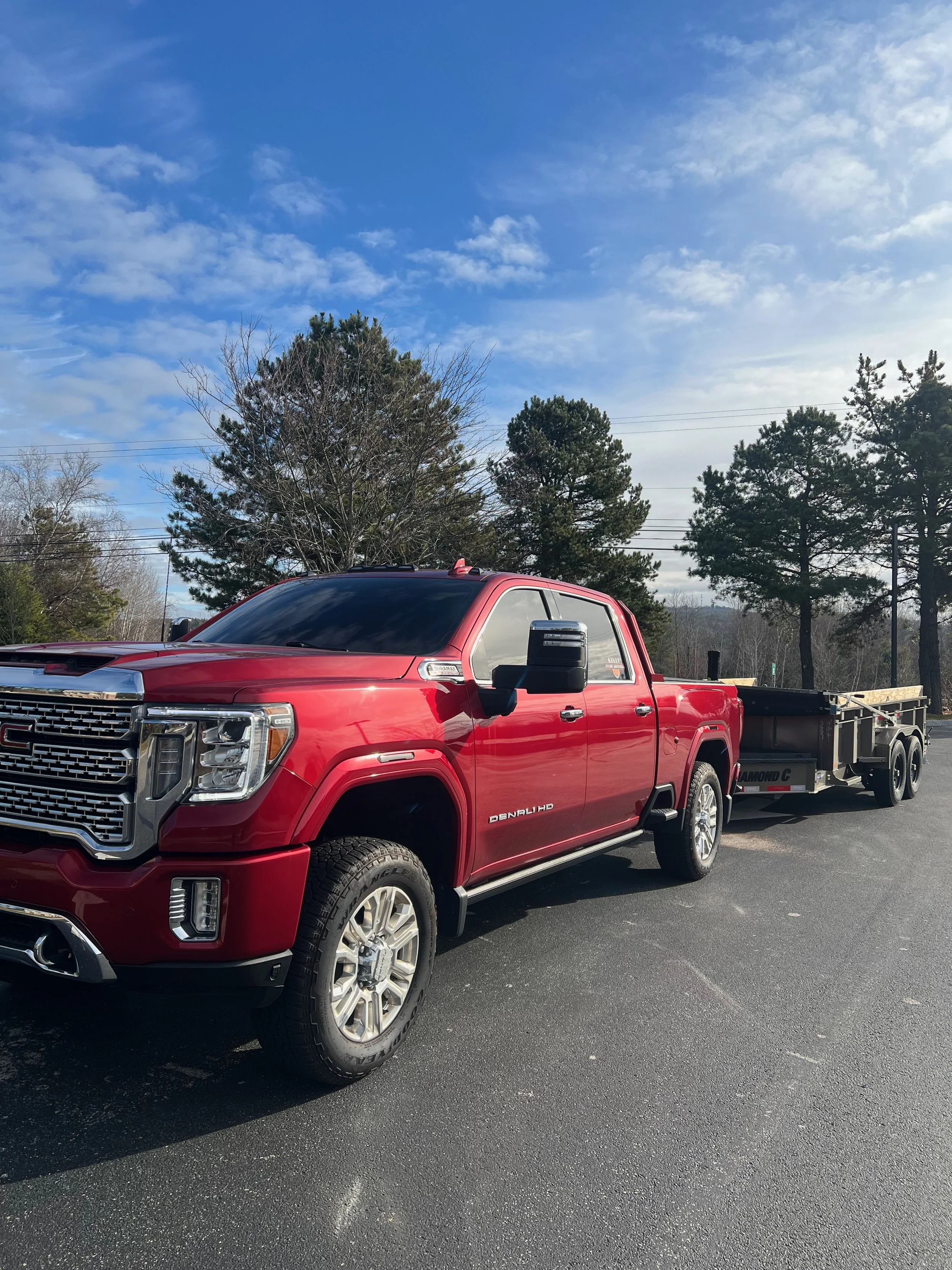 Red GMC Denali pickup truck with a trailer attached, parked on a paved lot with trees and blue sky in the background.