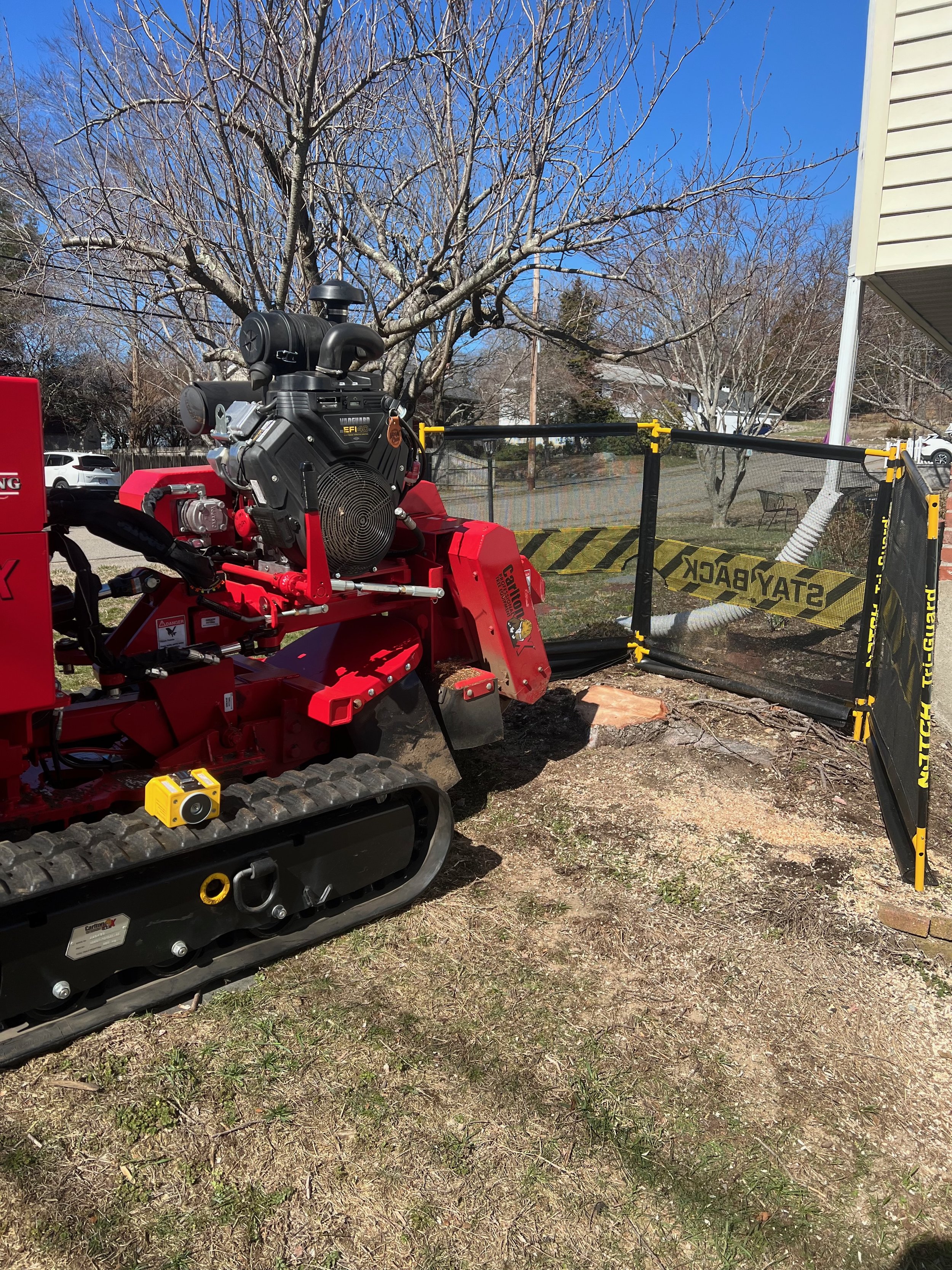 A small tracked skid steer loader with a black and red body is cutting a circular section of a tree stump. The tree has no leaves, and there is a wooded area, house, and cars in the background. The cutting area is protected by a black and yellow 'Sta