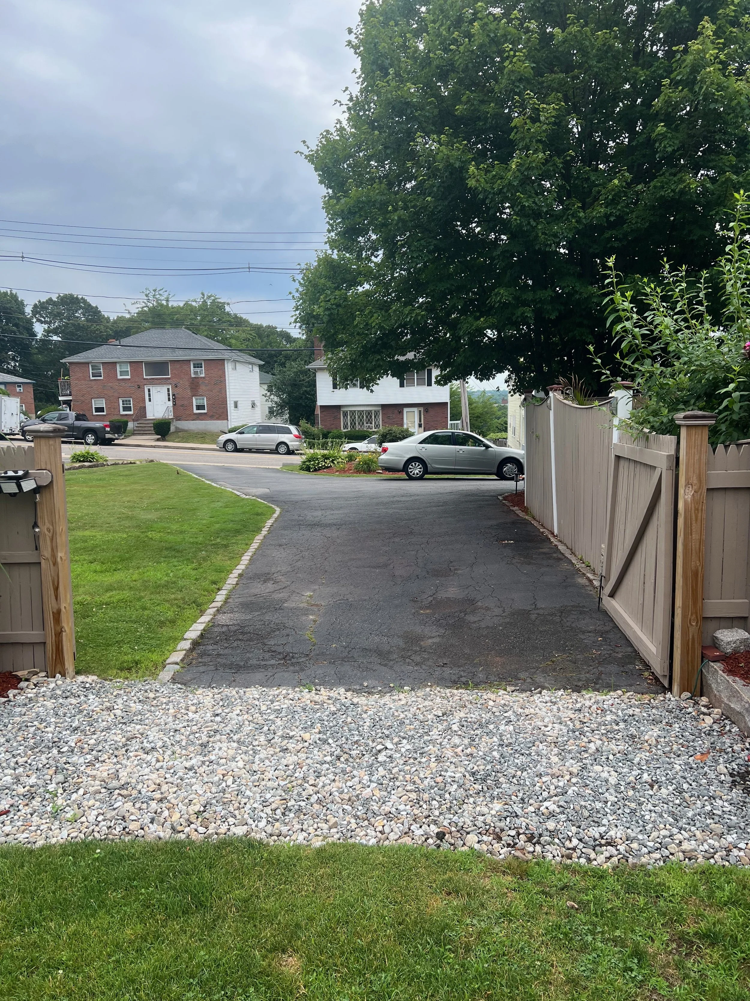 View of a residential driveway with a gravel area in the foreground, leading to an asphalt driveway with a gray car parked, bordered by a wooden fence and green lawn, with houses and trees in the background on a cloudy day.