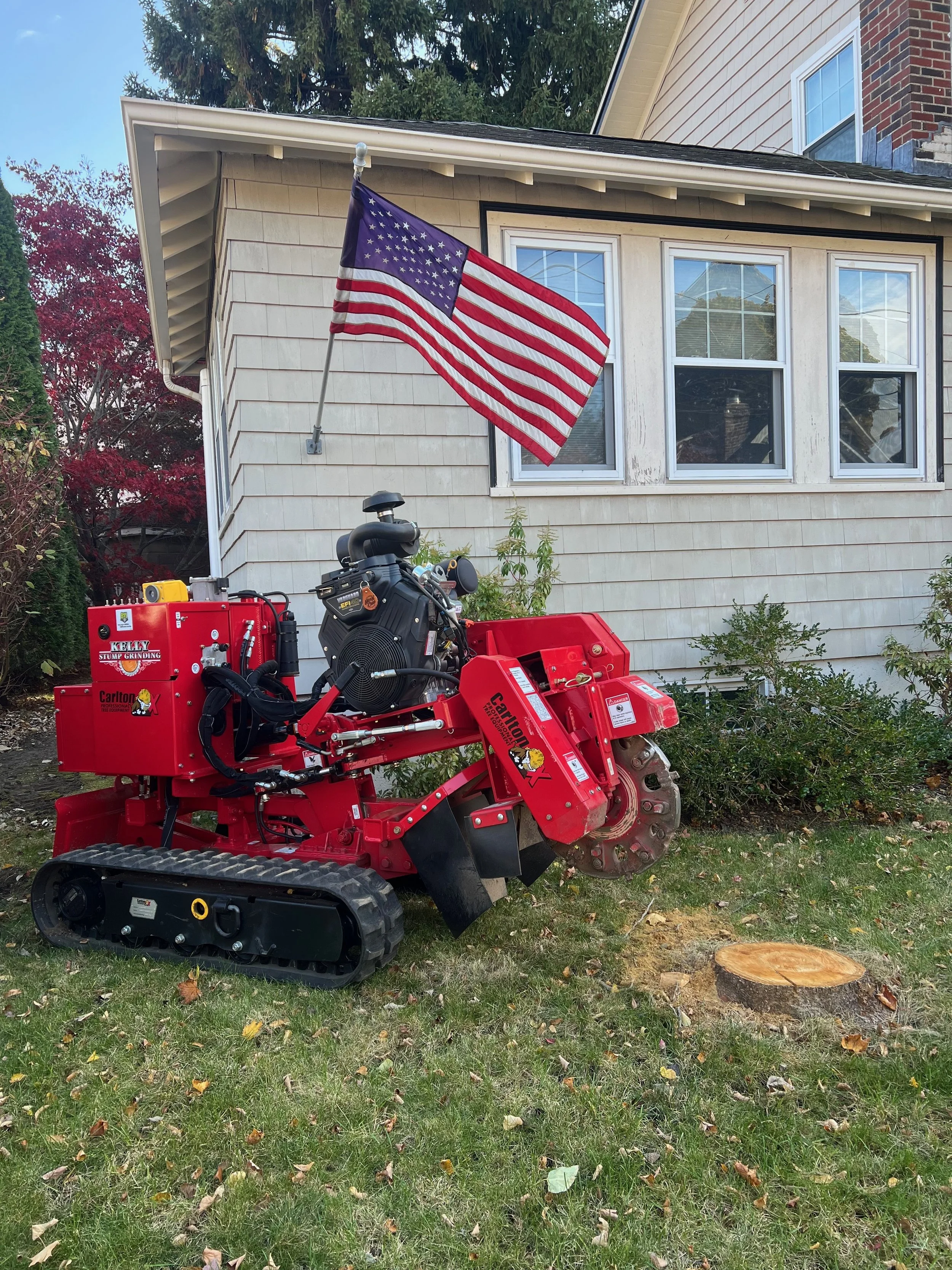 A red stump grinder machine with Caterpillar tracks, positioned on a lawn in front of a house with beige siding, white trim, and three windows. An American flag is attached to the house, waving in the wind, with trees and a clear sky in the backgroun