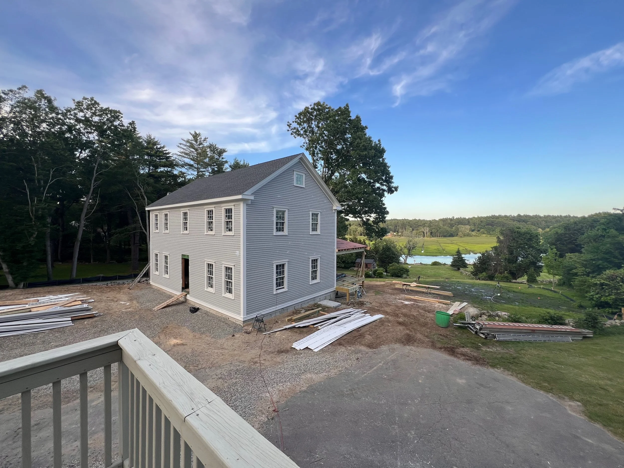 Under construction house with gray siding and multiple windows, construction materials on the ground, trees and a lake in the background, clear blue sky overhead.