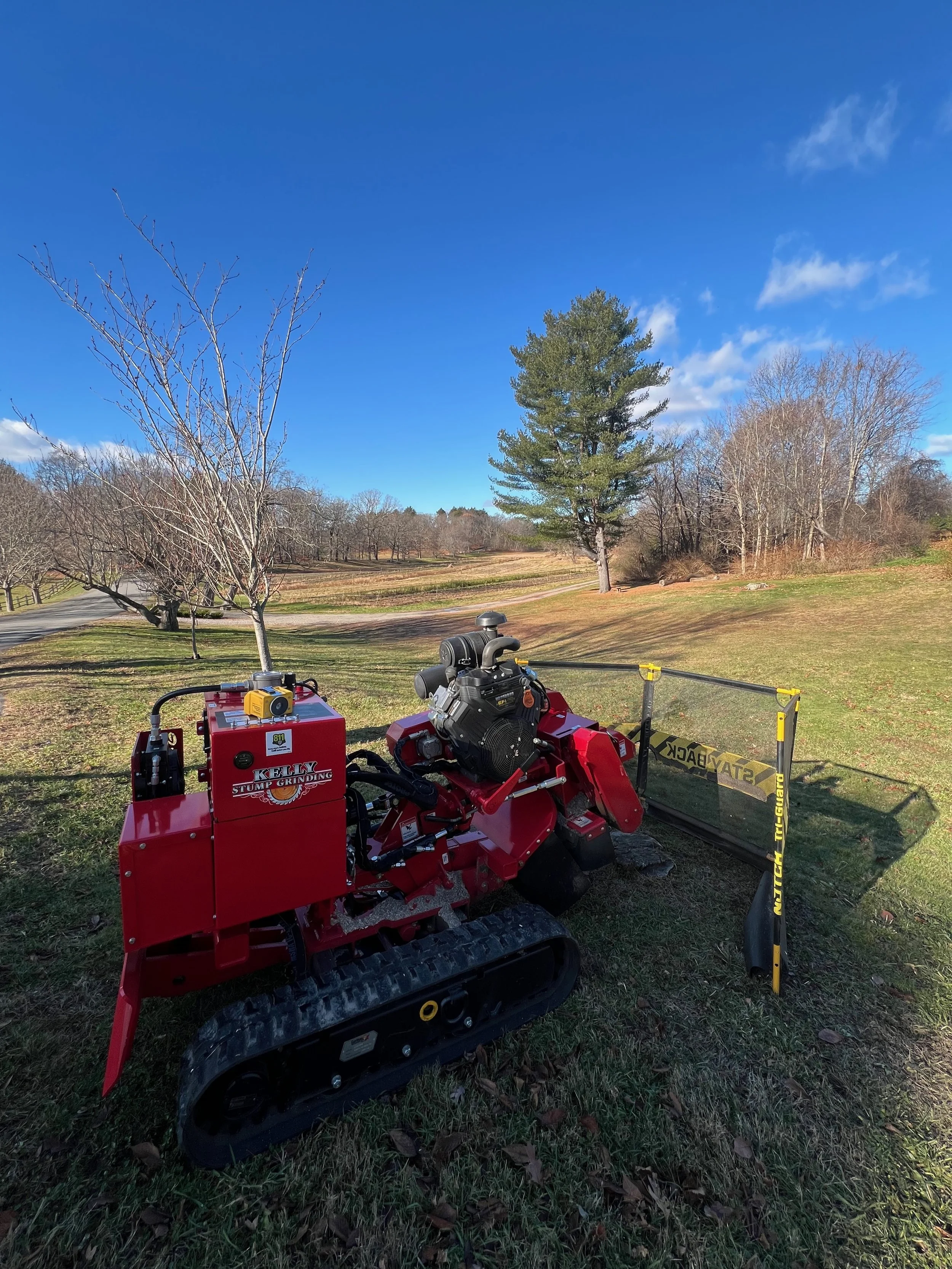 A red tracked stump grinder with black tracks and a yellow stump grinder attached, situated on a grassy area outdoors with trees and a clear blue sky in the background.