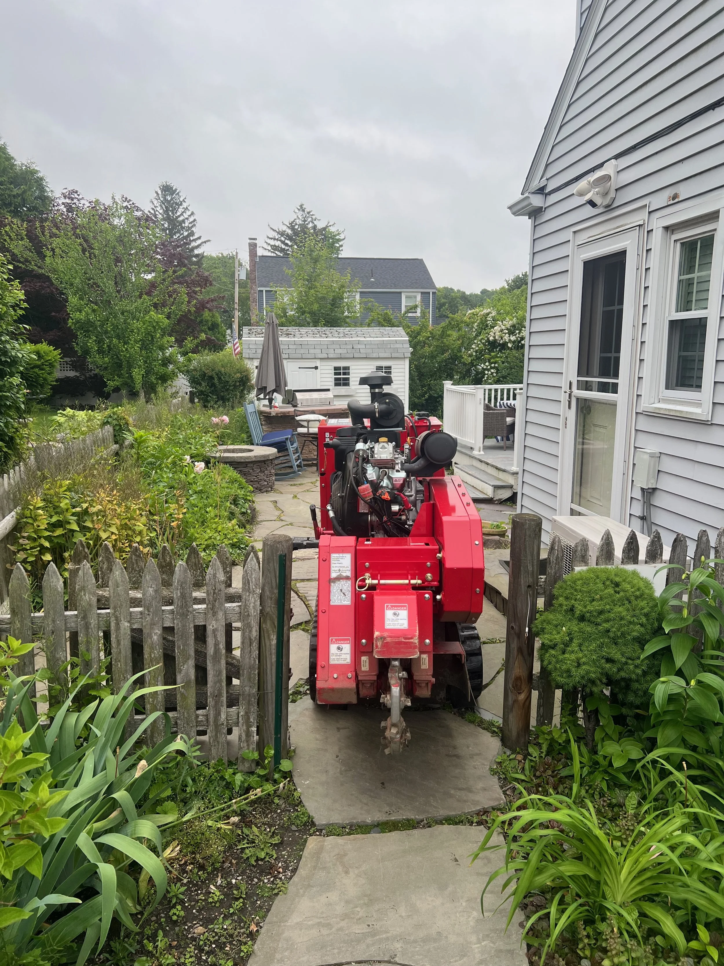 A red mini excavator parked on a stone pathway in a backyard surrounded by greenery. There are garden beds, a white house with a sliding door, and outdoor furniture in the background.