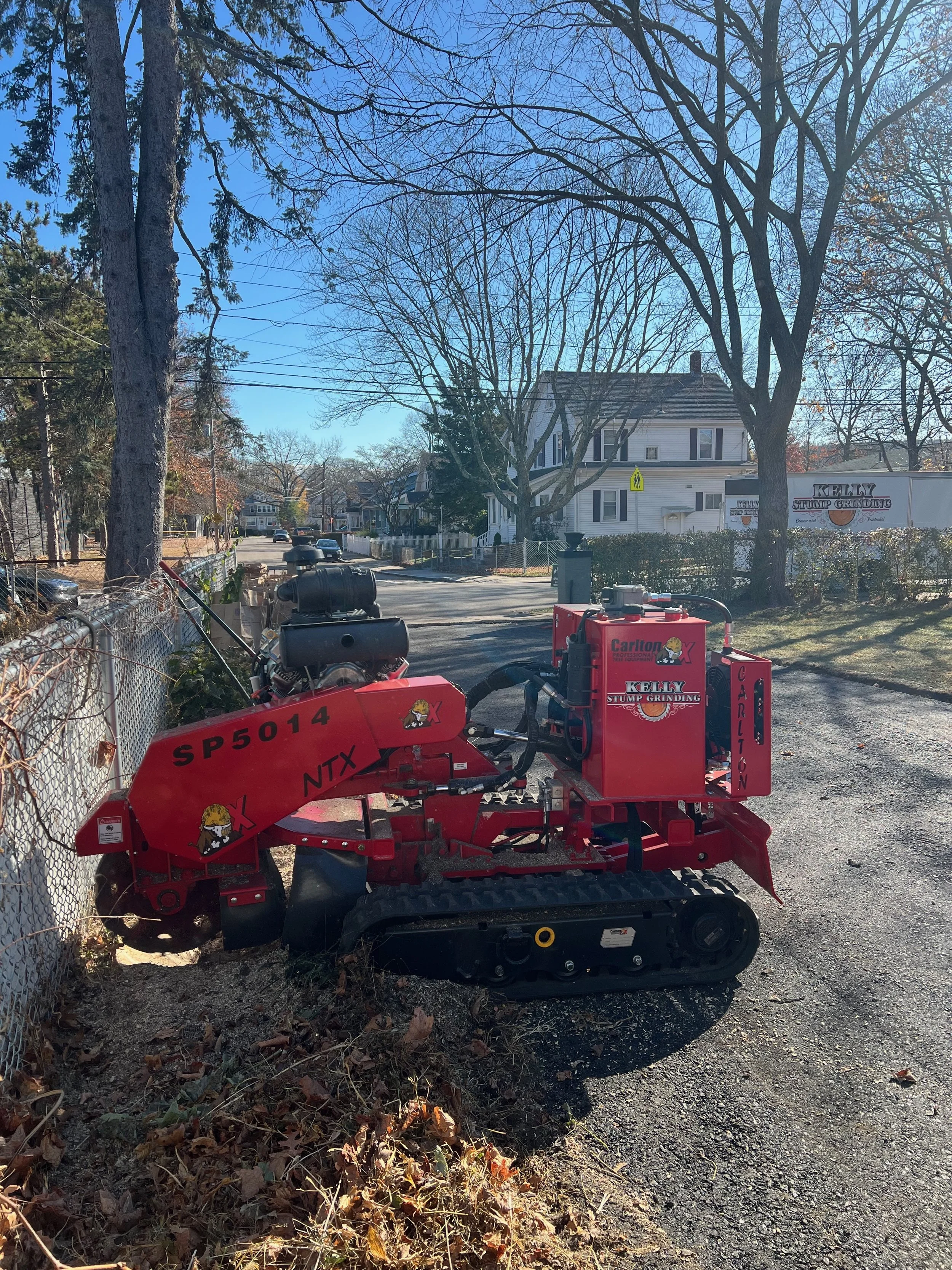 A small red tracked stump grinder with the words Kelly Stump Grinding and Carlton on it, positioned on a sidewalk next to a chain-link fence, with houses and trees in the background on a clear sunny day.
