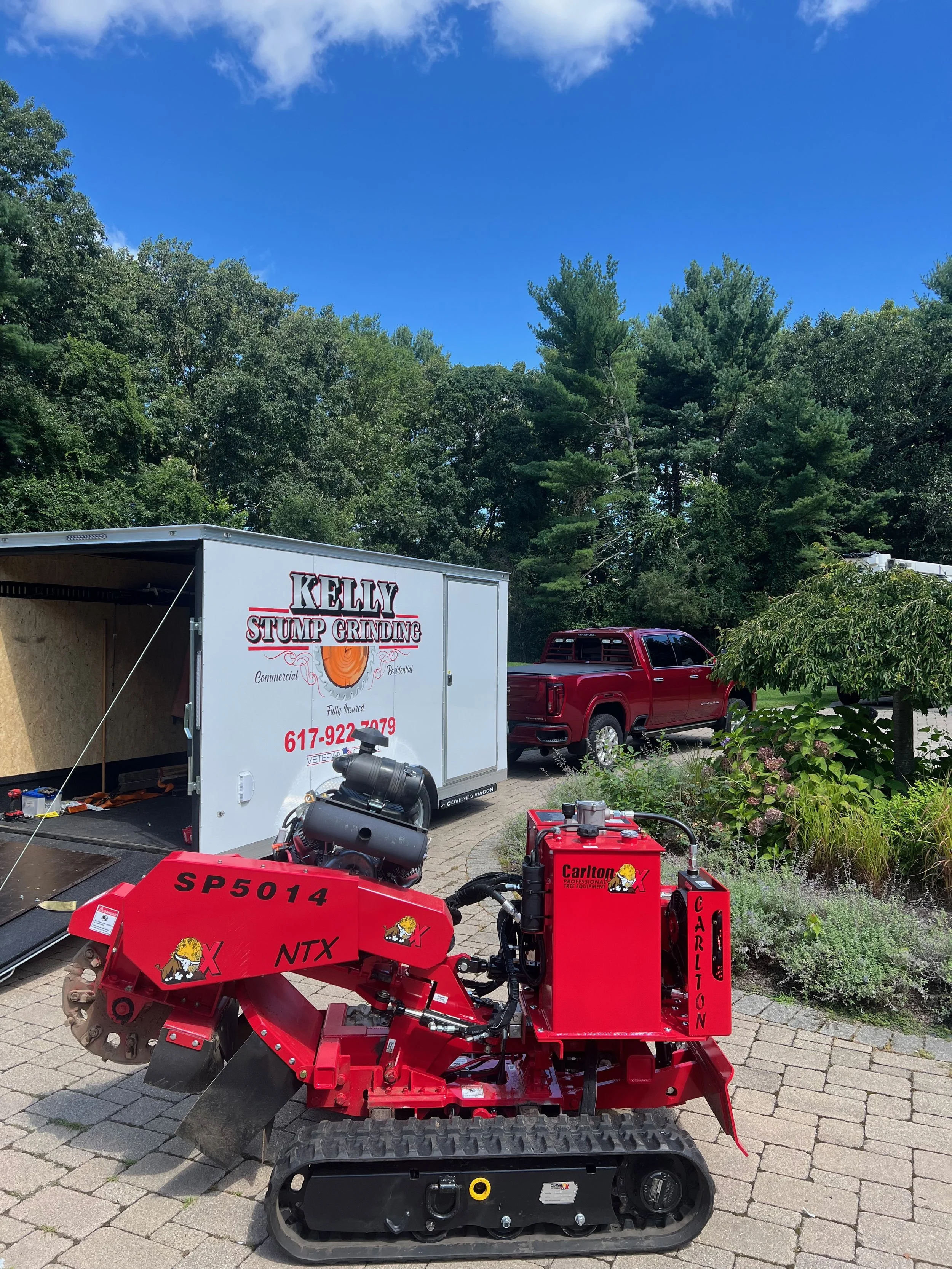 Red tracked stump grinder with black tracks, parked on a brick driveway, with a white trailer labeled 'Kelly Stump Grinding' and a red pickup truck in the background, surrounded by green trees and bushes.