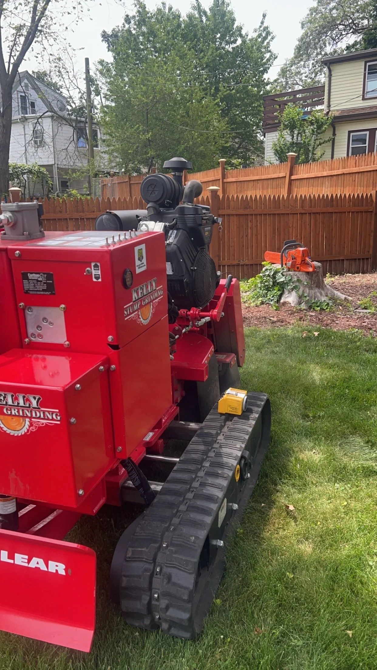 A small red tracked stump grinding machine on a lawn, with houses, trees, and a wooden fence in the background.
