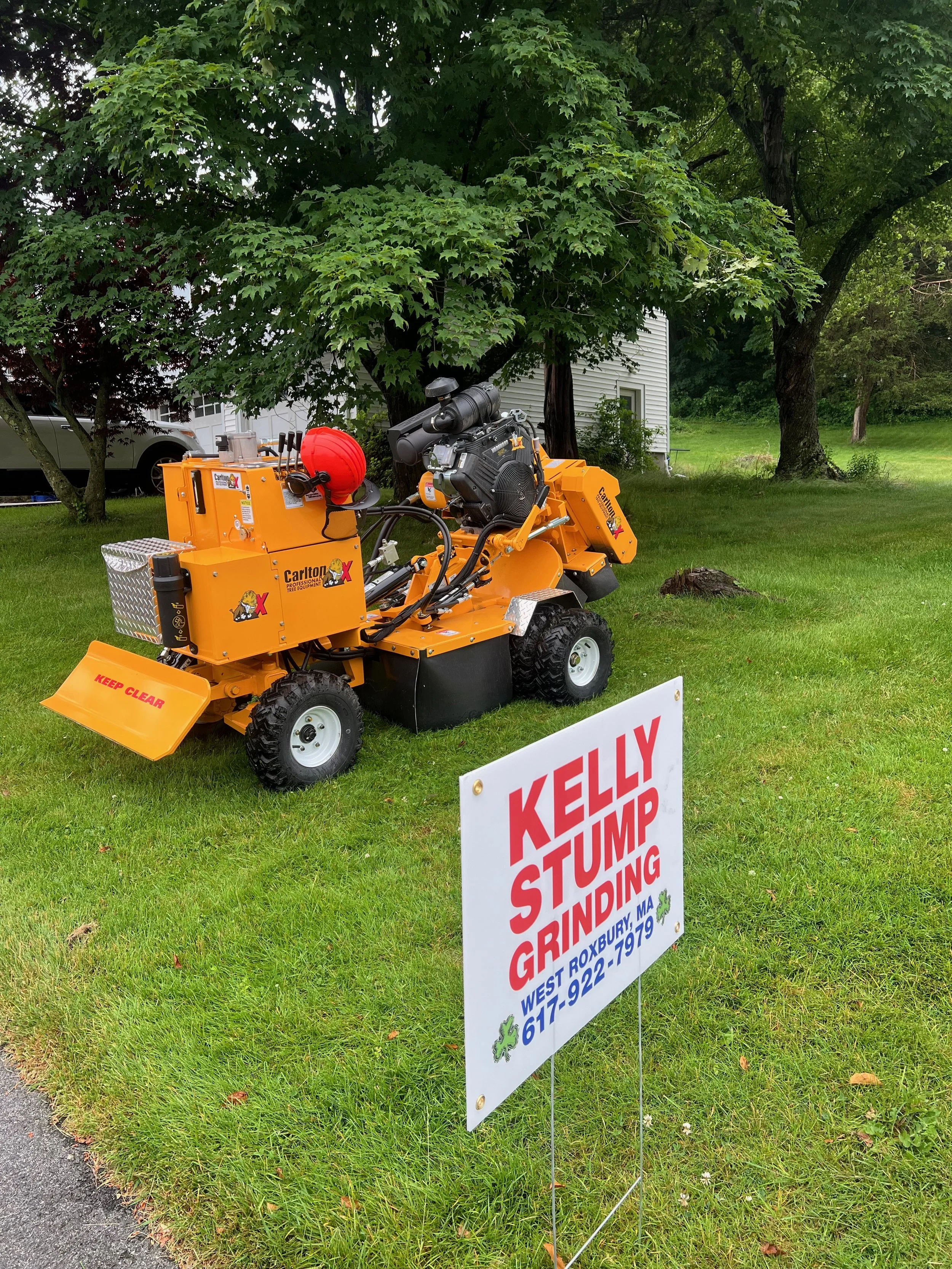 A piece of grinding equipment from Kelly Stump Grinding is parked on a grassy lawn with trees in the background and a sign in the foreground reading "Kelly Stump Grinding, West Roxbury, MA, 617-922-7979".