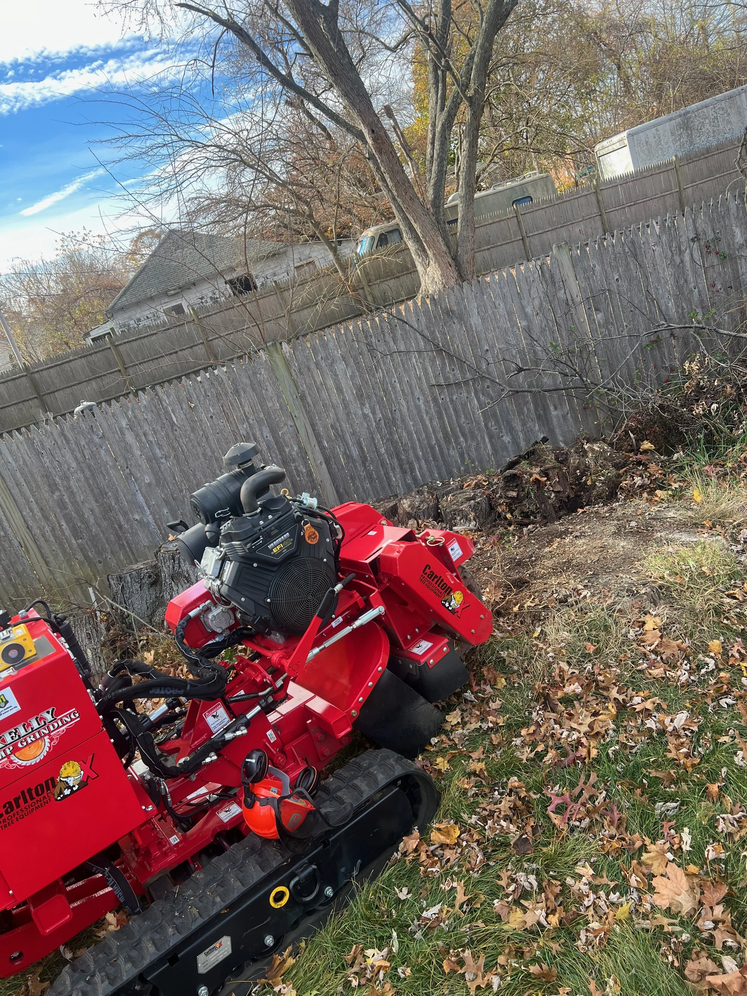 A red tree stump grinder with the brand name Carlton X in front of a wooden fence, autumn leaves on the ground, and a house with trees and a cloudy sky in the background.