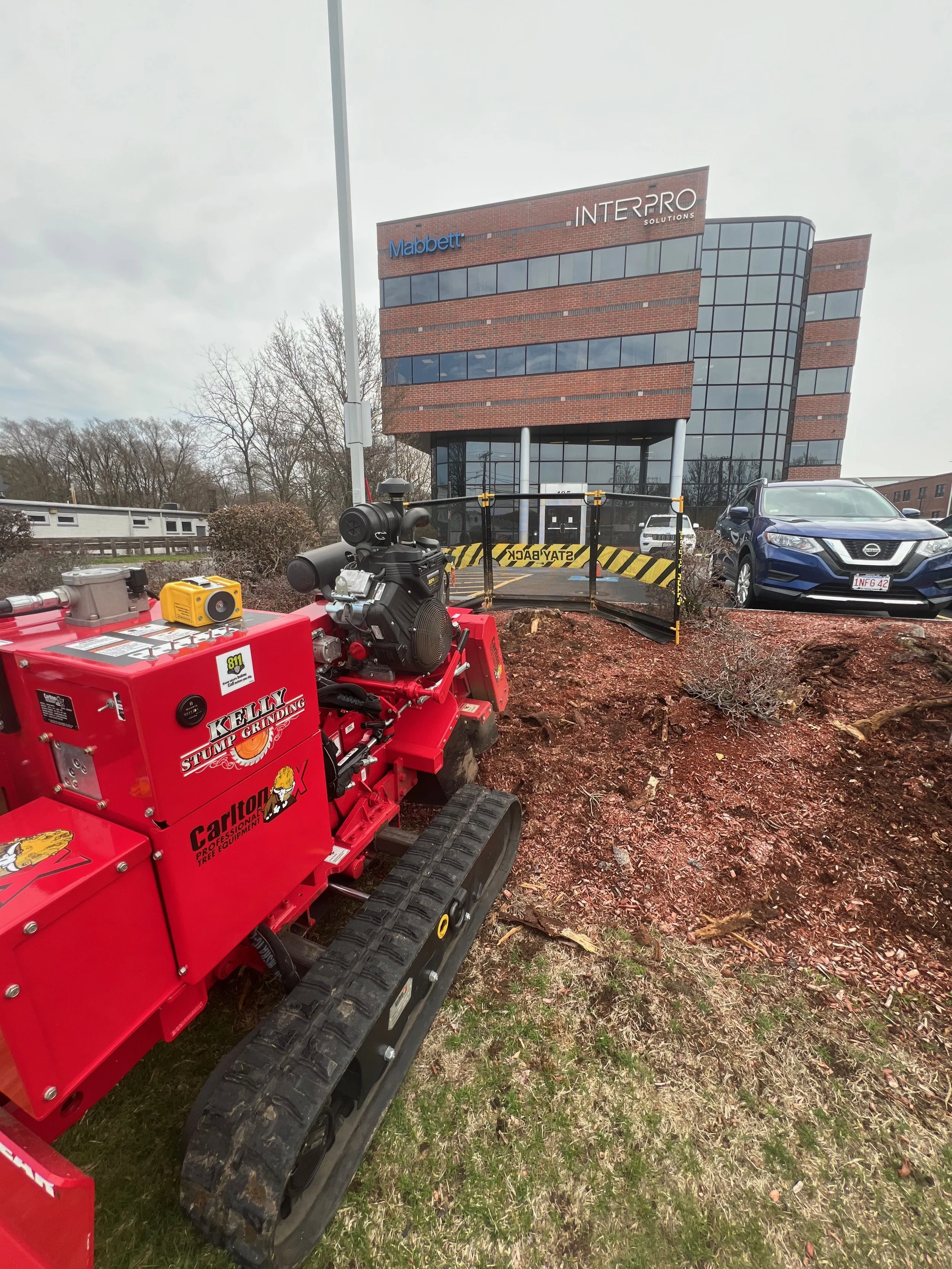 Construction bidirectional excavator with a red body and rubber tracks parked on a patch of grass in front of a modern office building with glass windows and brick exterior.