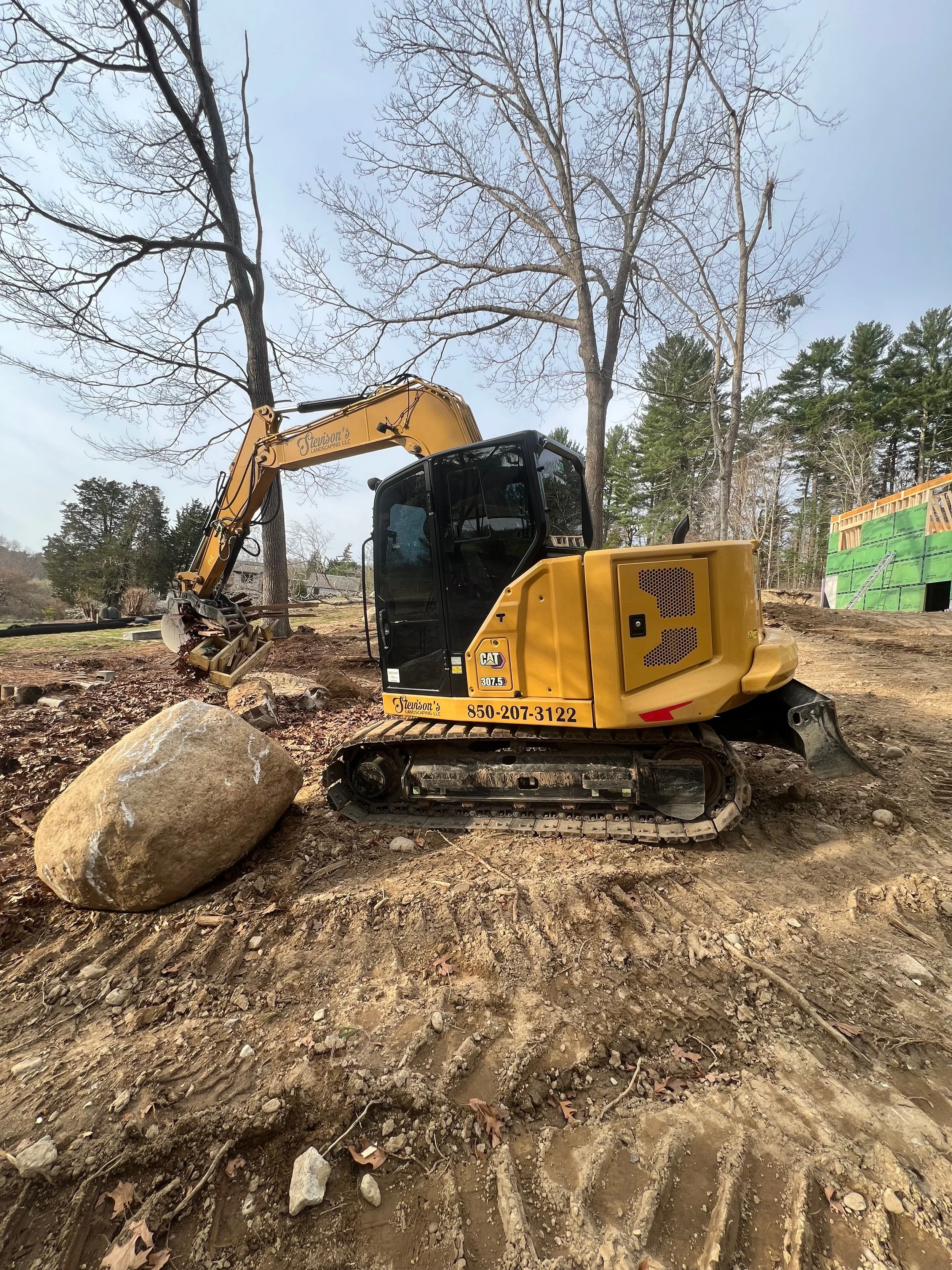 A yellow mini excavator with black tracks digging at a construction site, with a large boulder in front of it, and leafless trees and a green construction wall in the background.