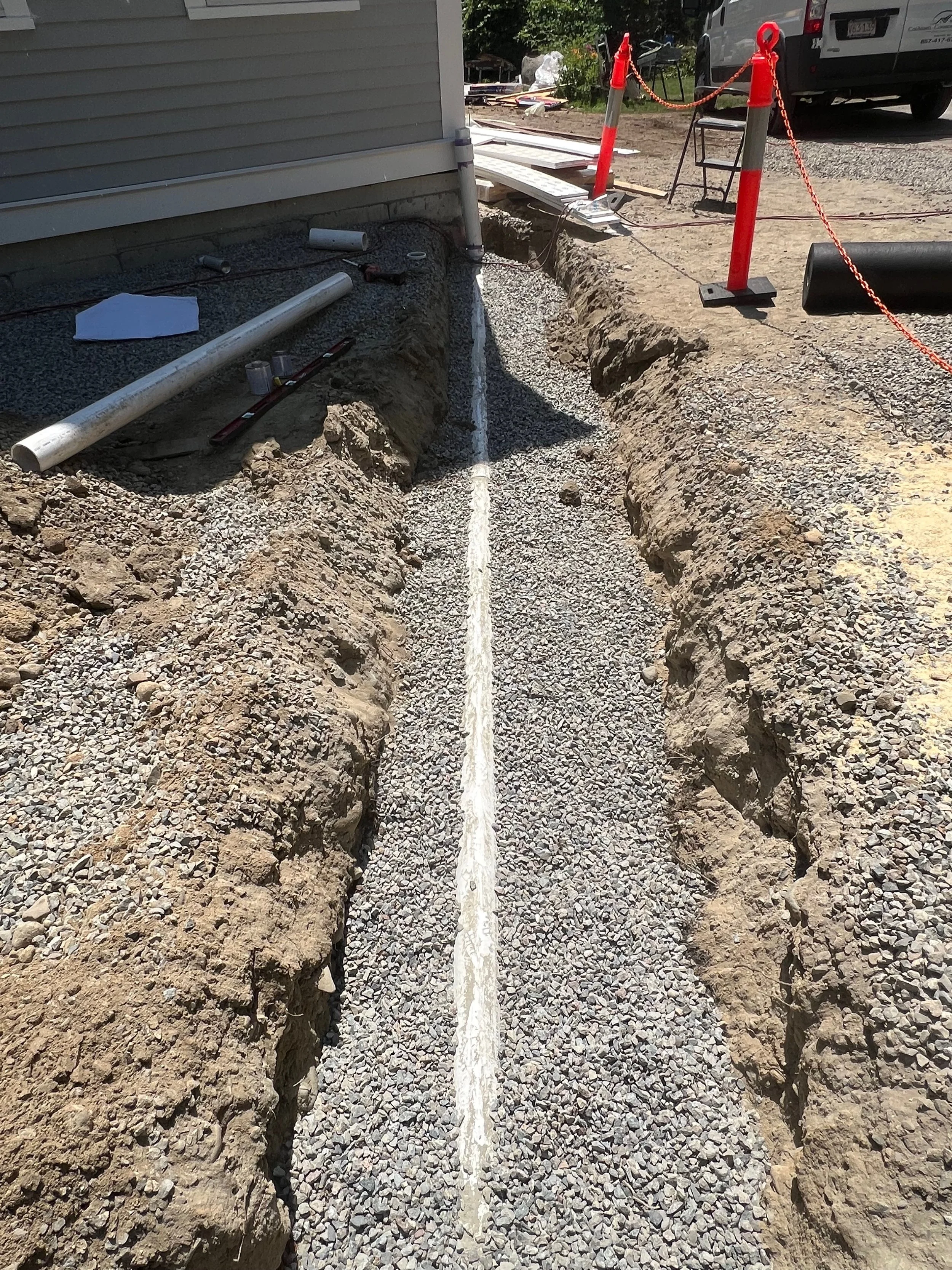 Construction site with a trench for underground utility pipes, with PVC pipes and tools on the side, and traffic cones and barriers in the background.