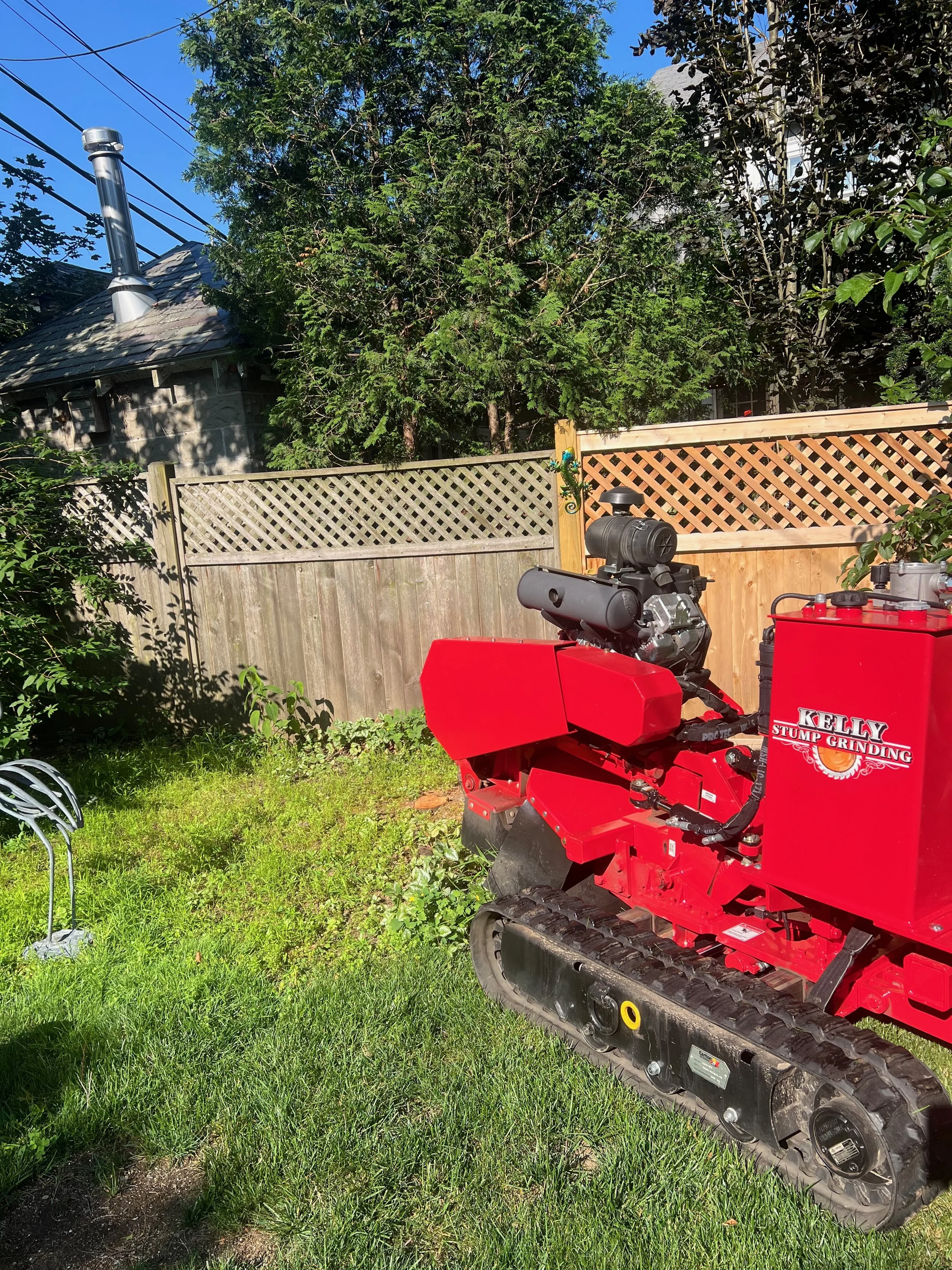 A red Kelly stump grinding machine on a patch of grass in a backyard, with a wooden fence, trees, and a house with a chimney in the background.