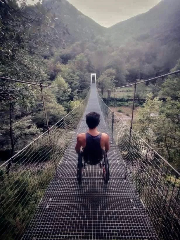Une personne en fauteuil roulant regarde un pont suspendu dans une vallée boisée, entourée de montagnes.