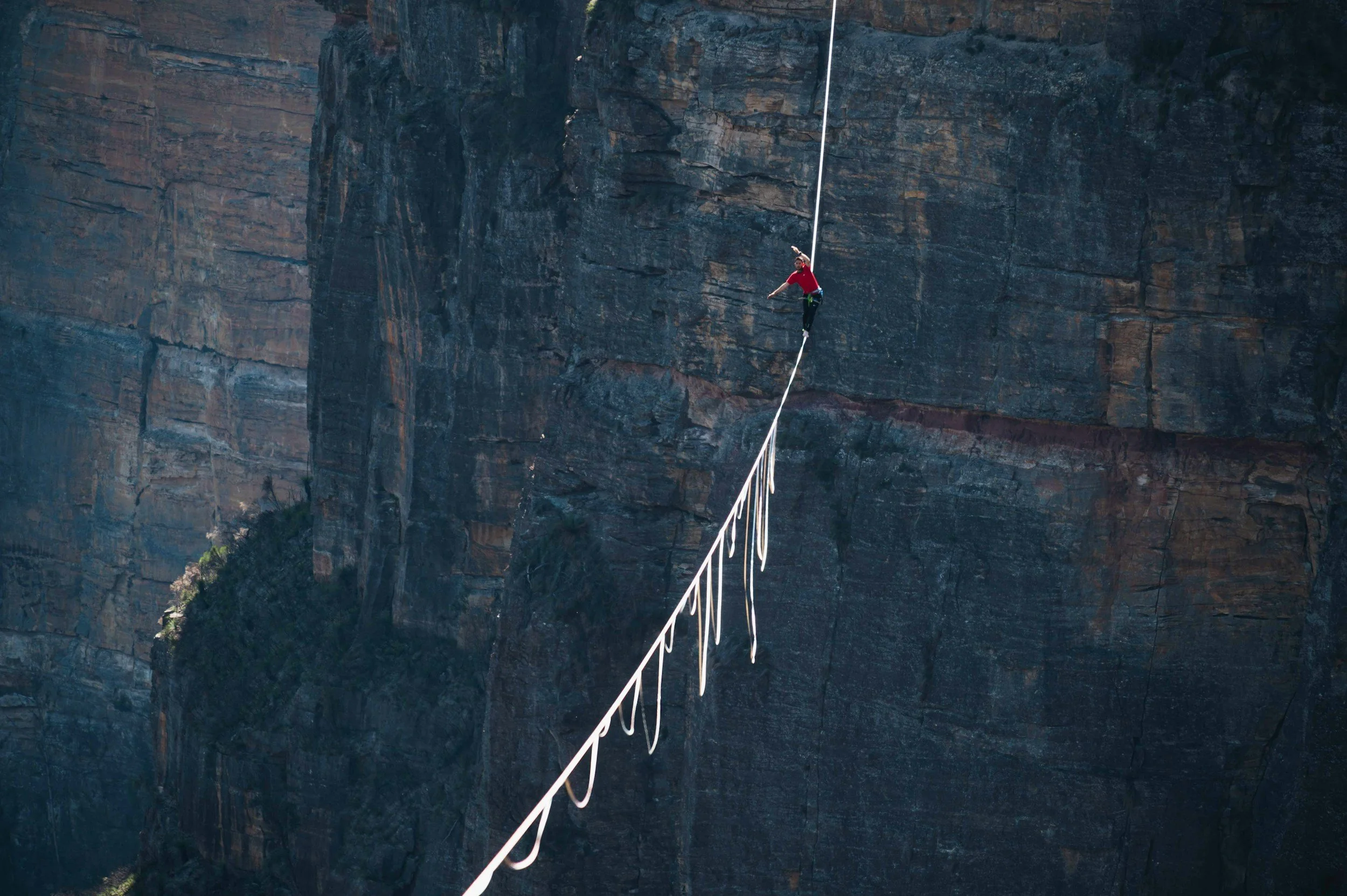 Une personne en t-shirt rouge marche sur une corde tendue à haute altitude au-dessus d'une falaise rocheuse.