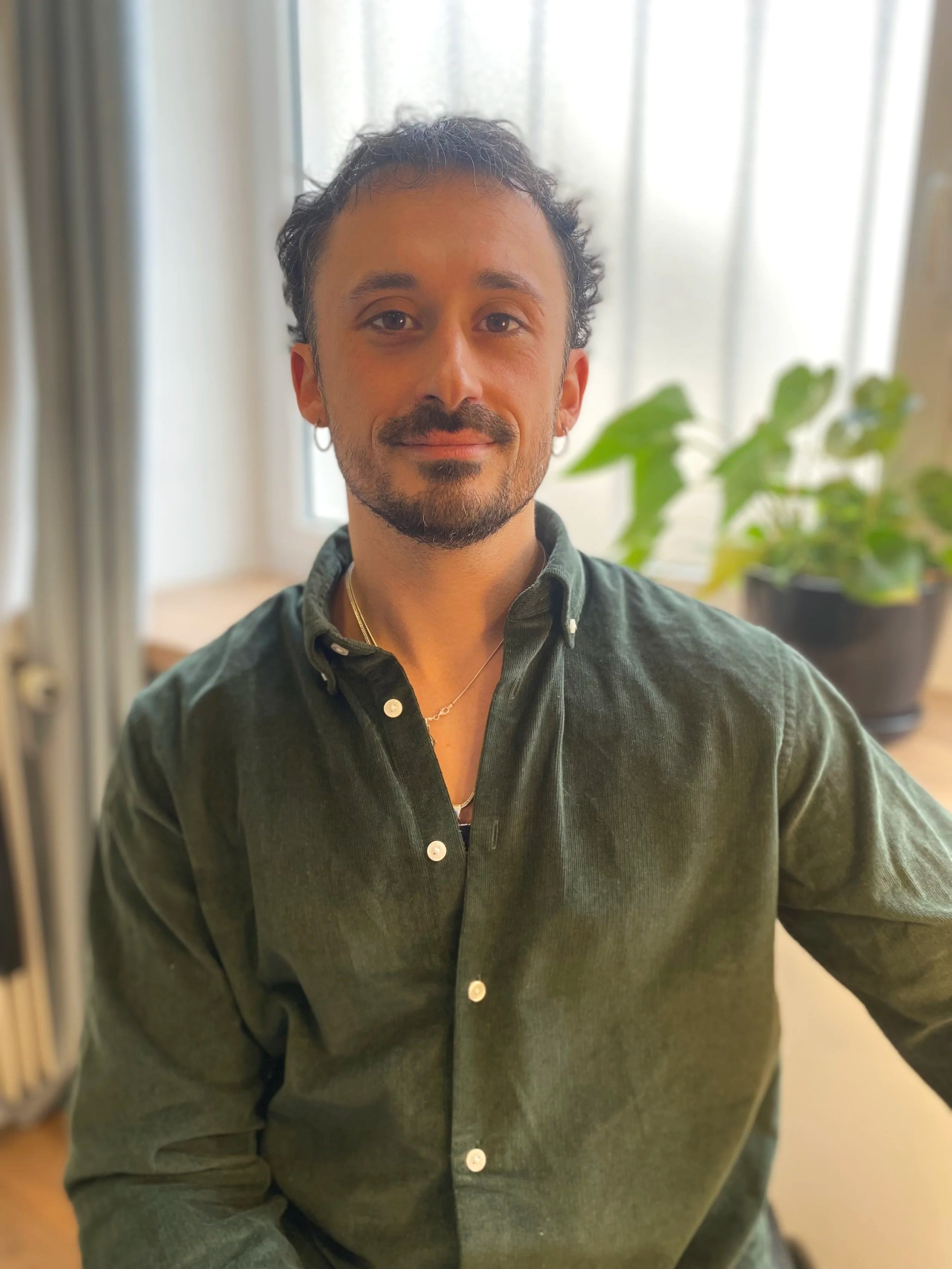 Un homme aux cheveux courts et bouclés, portant une chemise verte, sourire, à l'intérieur, avec une plante en pot derrière lui.