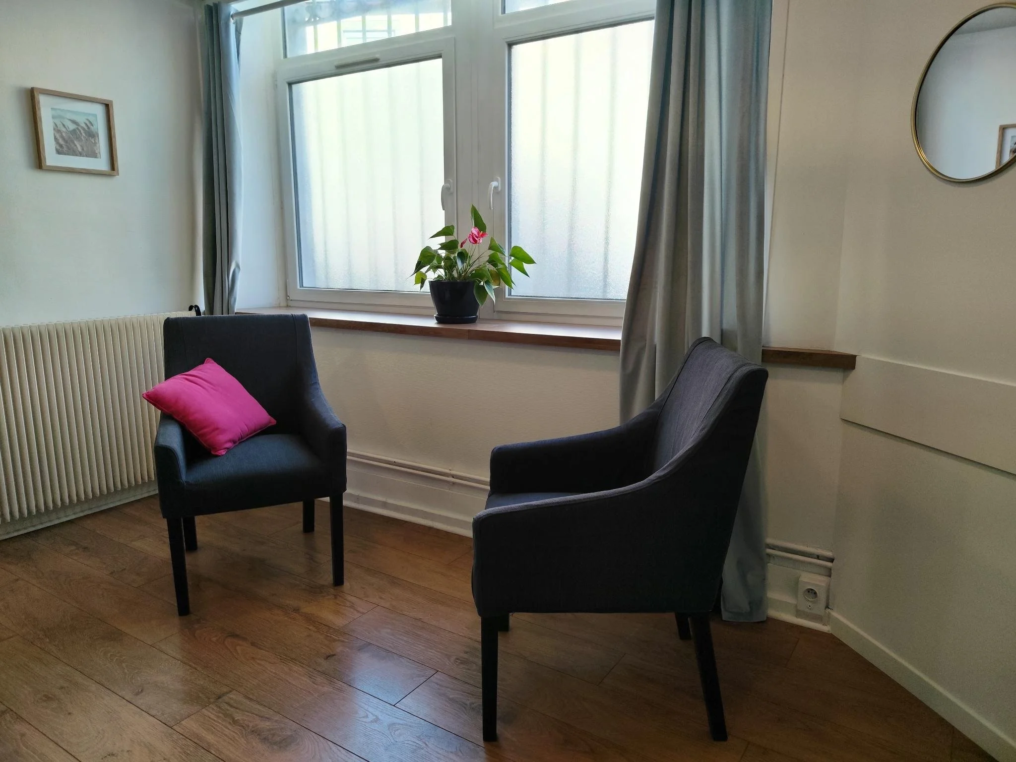 Une pièce lumineuse avec deux fauteuils gris, l'un avec un coussin rose, une fenêtre avec rideaux gris et une plante en pot sur le rebord. Il y a aussi un miroir rond et une peinture au mur.