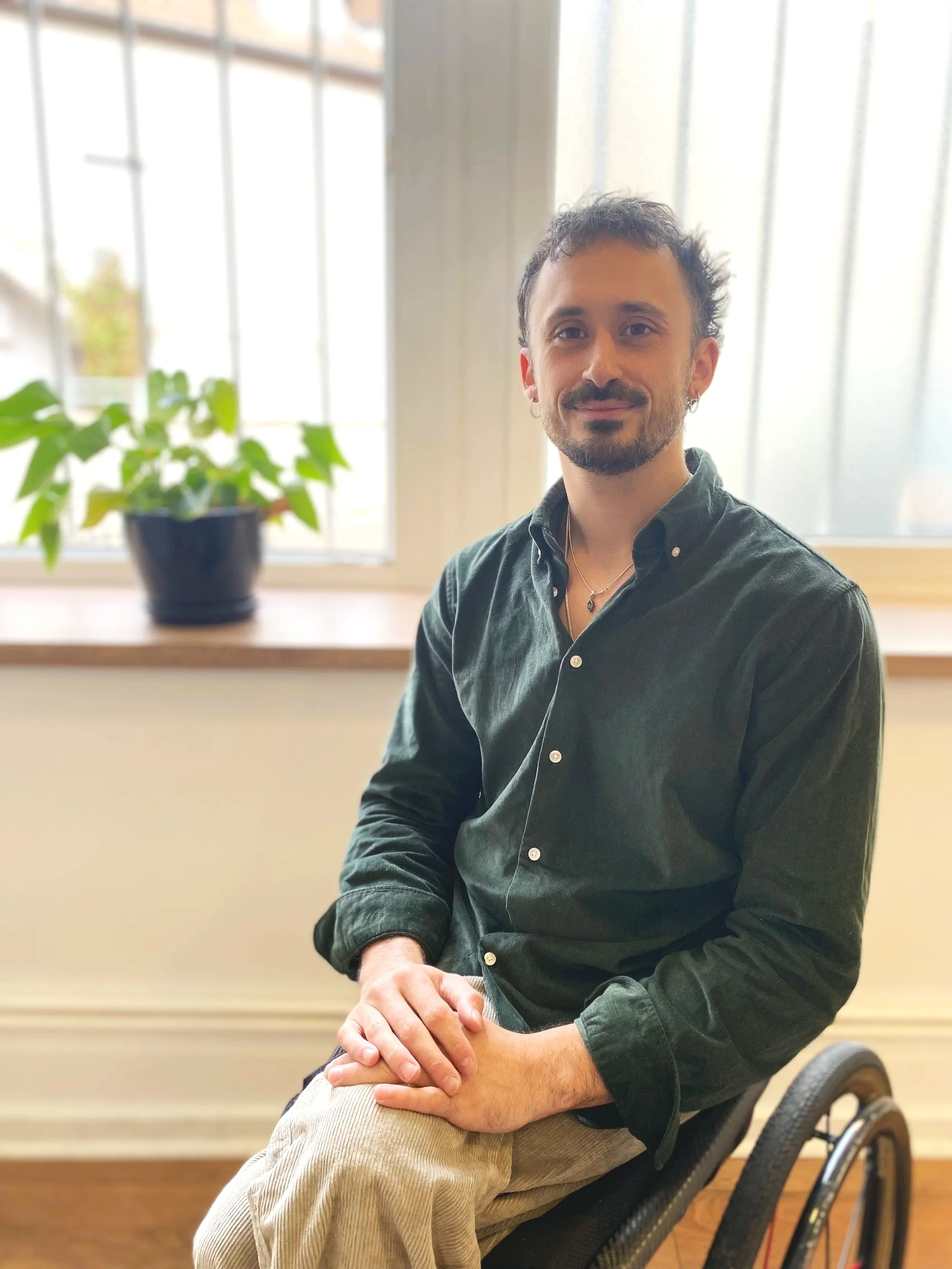 Homme en fauteuil roulant, portant une chemise vert foncé, assis à l'intérieur près d'une fenêtre avec une plante verte en pot en arrière-plan.