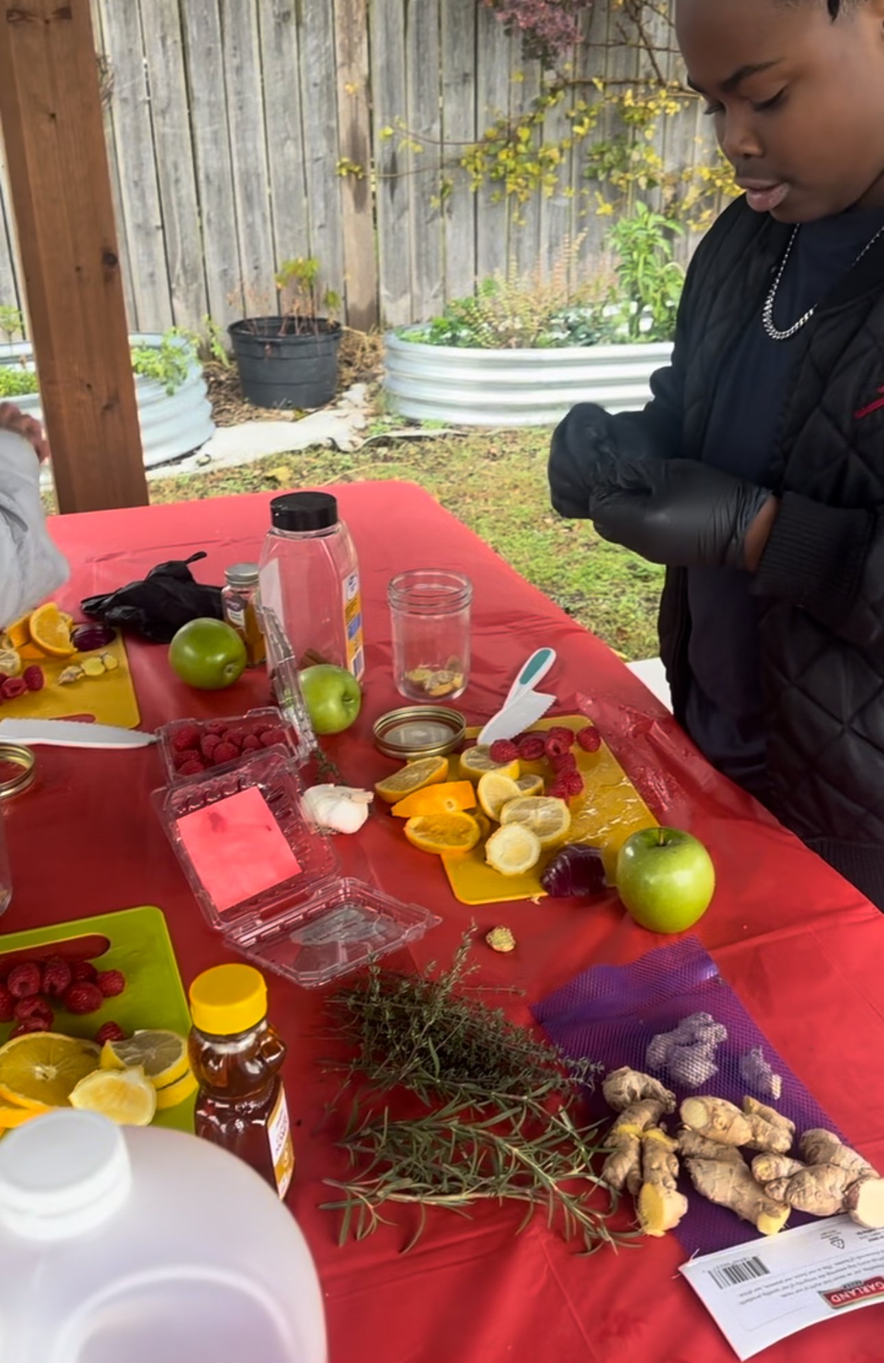 Outdoor table with assorted chopped fruits like oranges, bananas, and berries, fresh herbs, and various jars and bottles, with a woman wearing gloves standing beside.