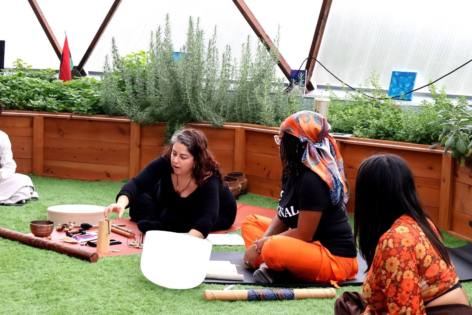 Three women sitting on the grass in a greenhouse, engaging in a spiritual or cultural activity. The woman on the left is reaching towards instruments and objects in front of her. The woman in the middle, wearing a colorful headscarf and glasses, is s
