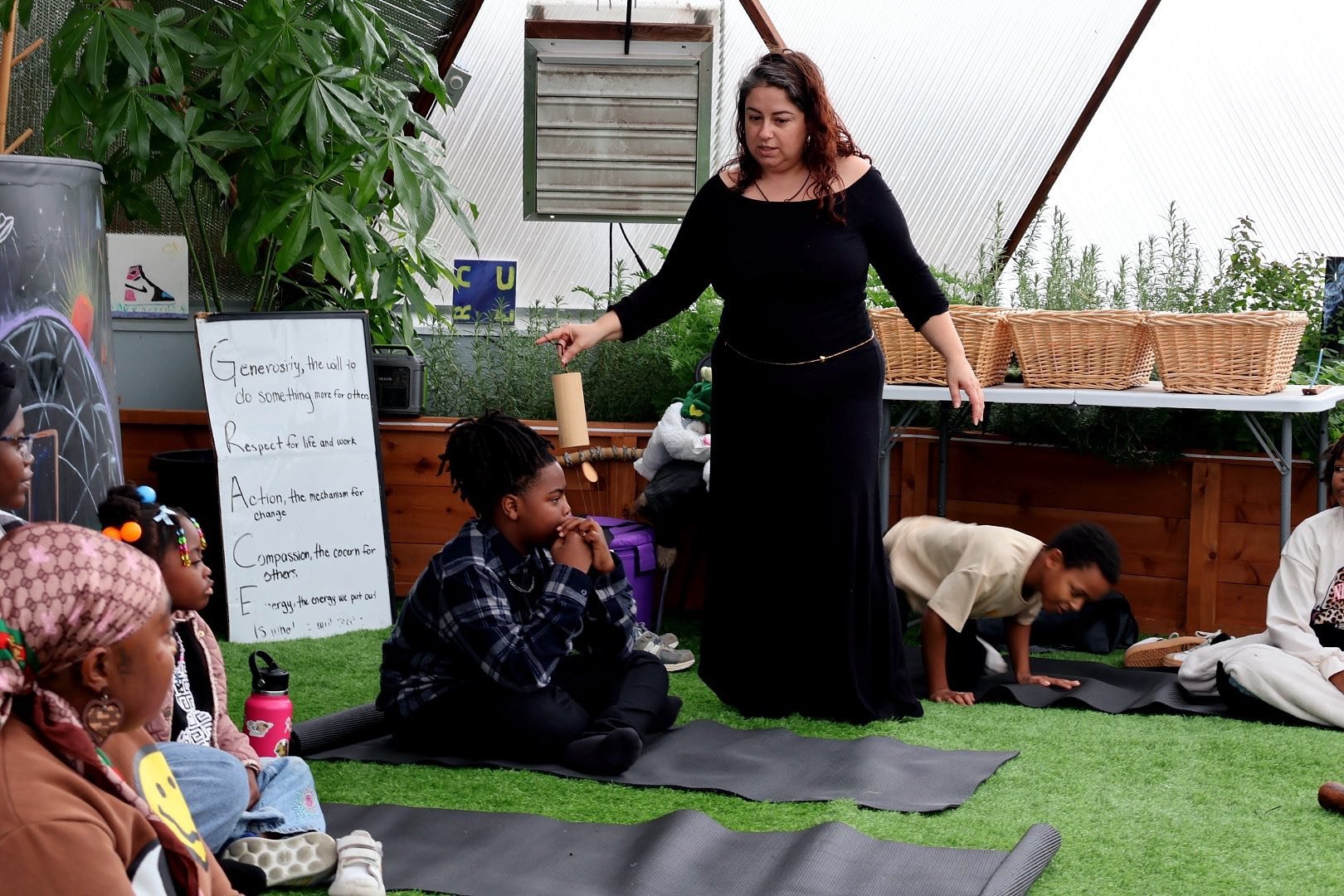 A woman leading a group meditation or yoga session with children sitting on yoga mats in a greenhouse or indoor garden setting, with a whiteboard in the background displaying a message about generosity.