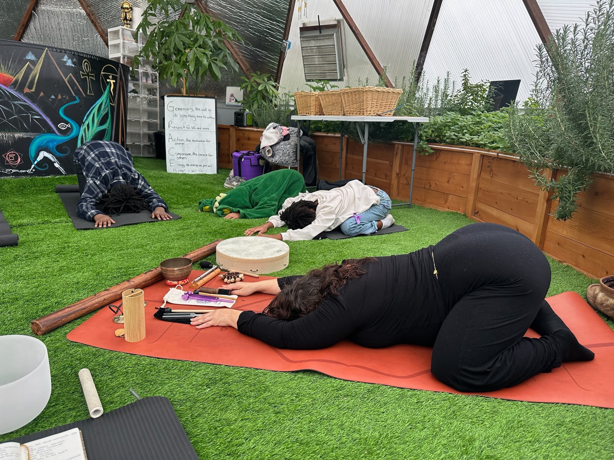 Four people participating in a yoga or meditation class in a greenhouse with plants lining the walls, yoga mats on the grass, and various meditation props nearby, including singing bowls and a drum.