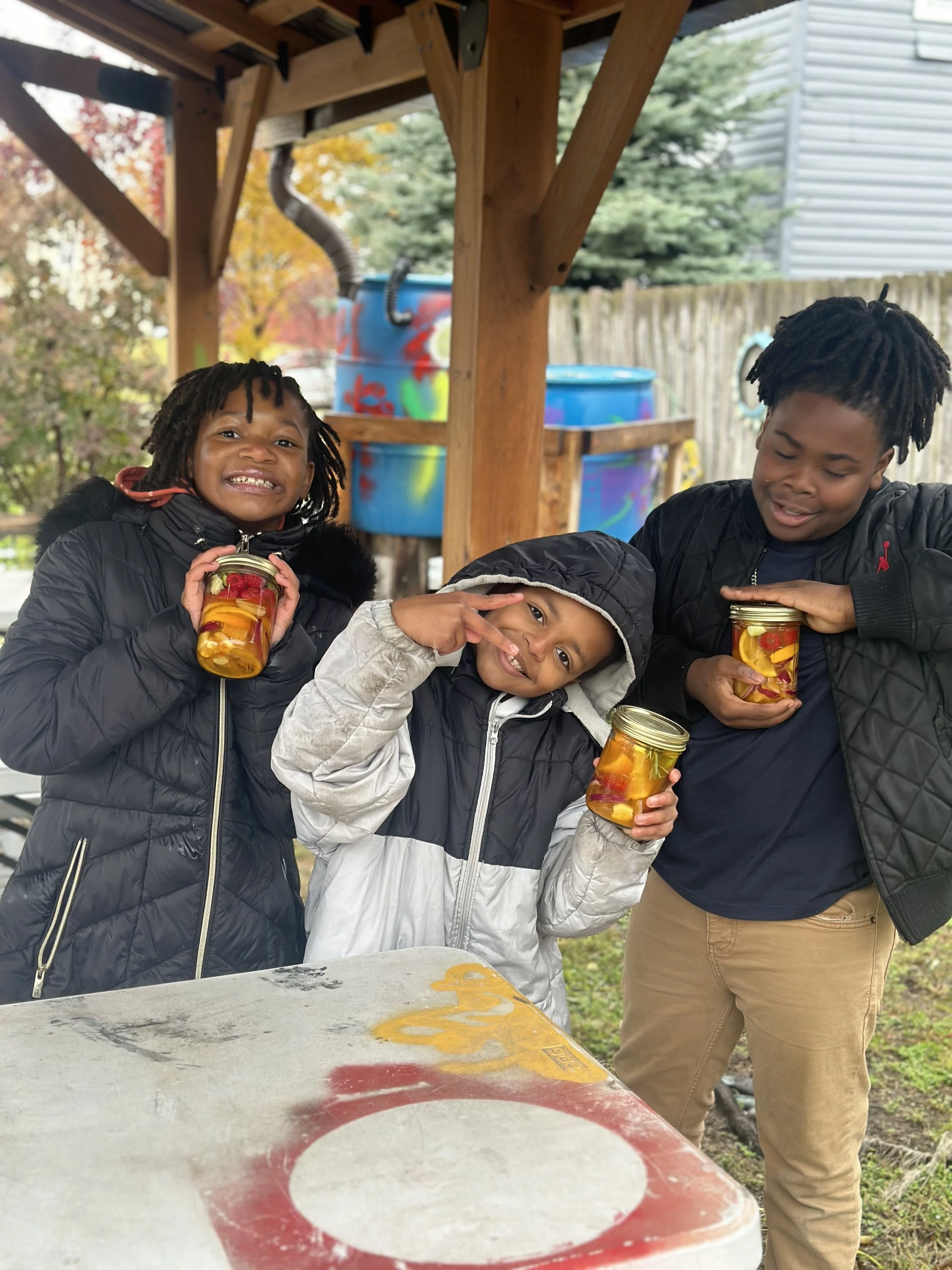 Three children standing under a wooden shelter, holding jars of fire cider smiling and making playful gestures, with a colorful outdoor background.