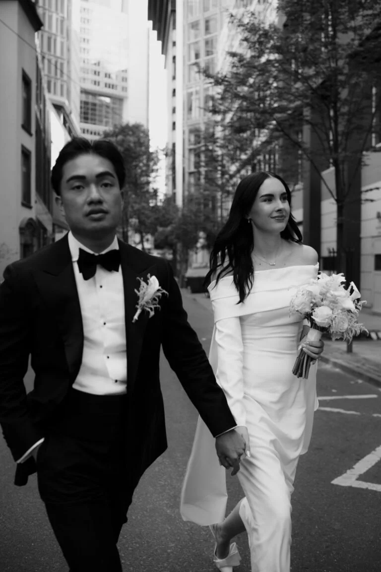 A black-and-white photograph of a newlywed couple holding hands and walking down a city street. The groom is dressed in a tuxedo with a bow tie and boutonniere, and the bride wears a white off-the-shoulder wedding dress and holds a bouquet of flowers.
