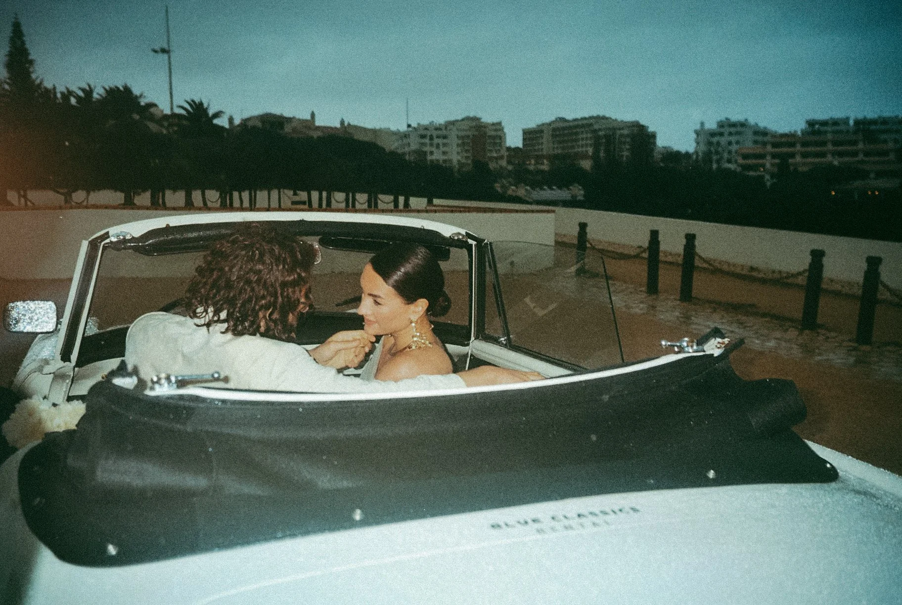 Two women are in a convertible car at dusk, with one leaning in to touch the other's face, both dressed elegantly.