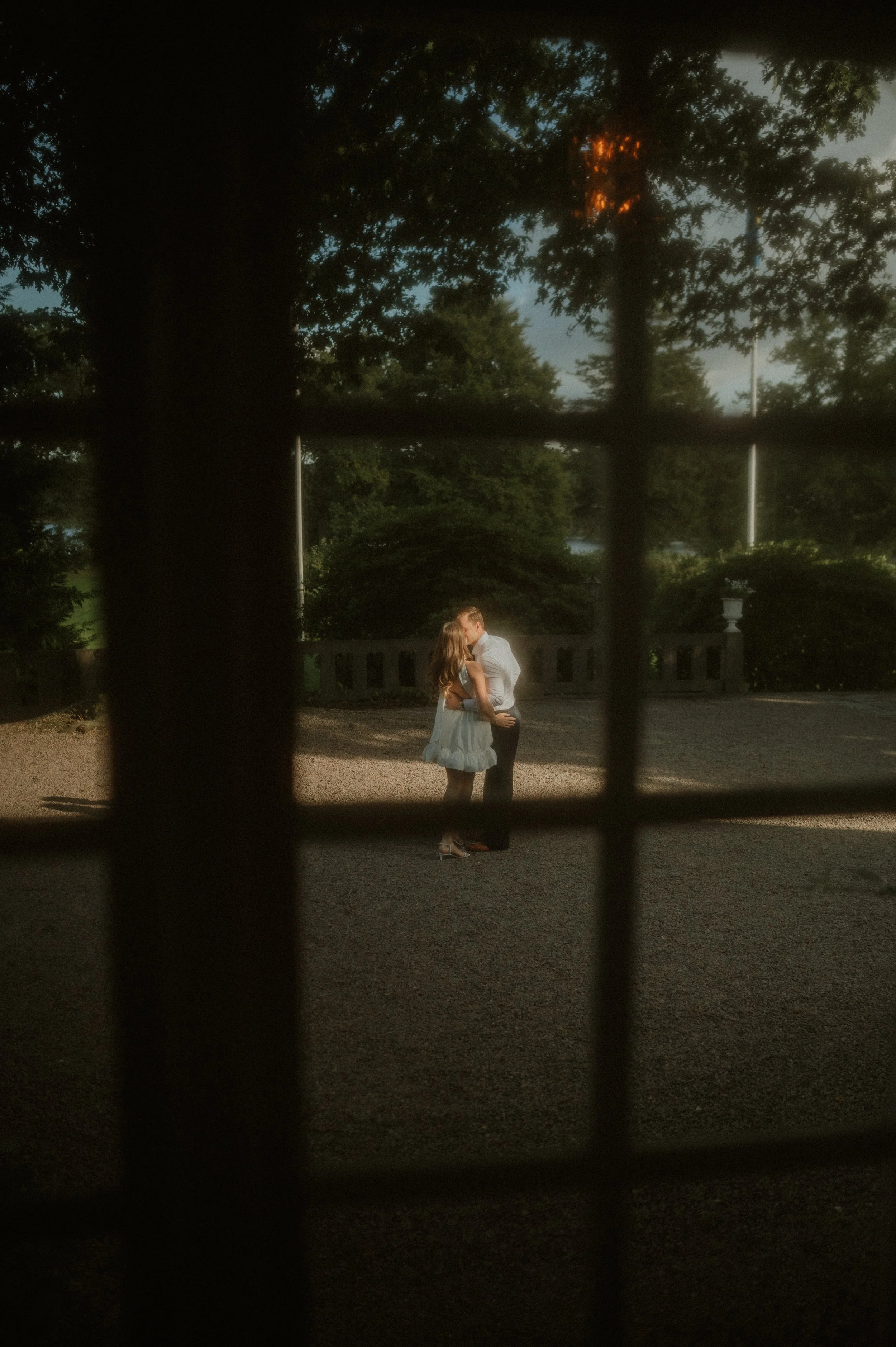 A couple kissing outdoors seen through window panes with a grid pattern, surrounded by greenery, trees, and a stone railing.