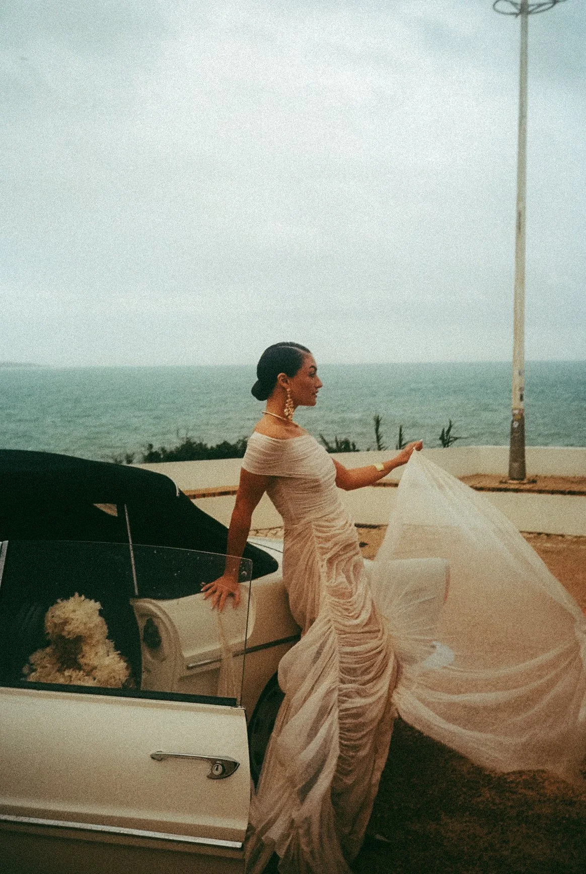 A woman in a flowing, elegant beige dress with a draped design, sitting on the door of a vintage white car with a flower bouquet inside, near the beach with the ocean and cloudy sky in the background.
