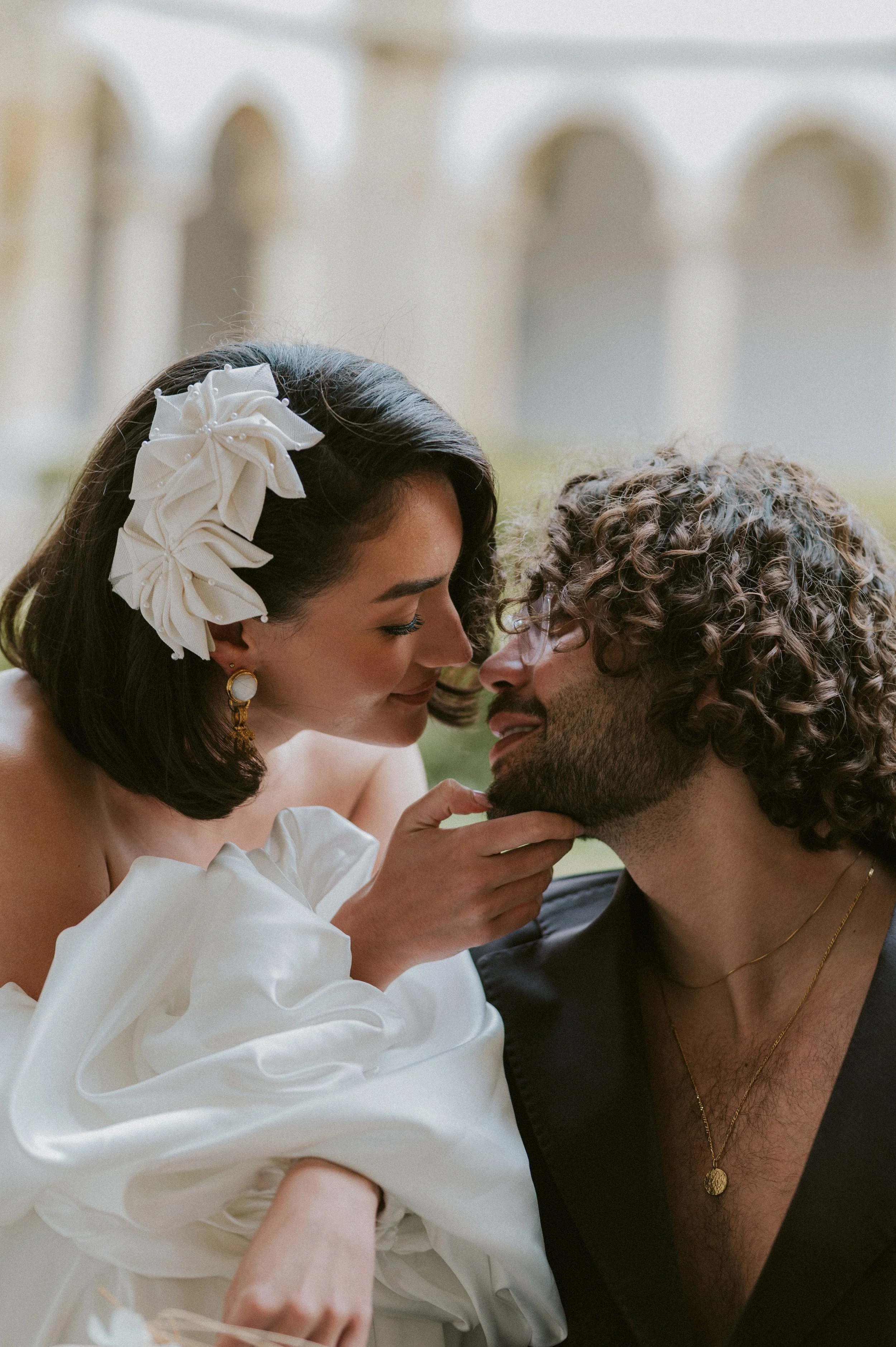 A couple sharing an intimate moment, with the woman gently touching the man's chin, both smiling and looking into each other's eyes, in an outdoor setting with classical architecture in the background.