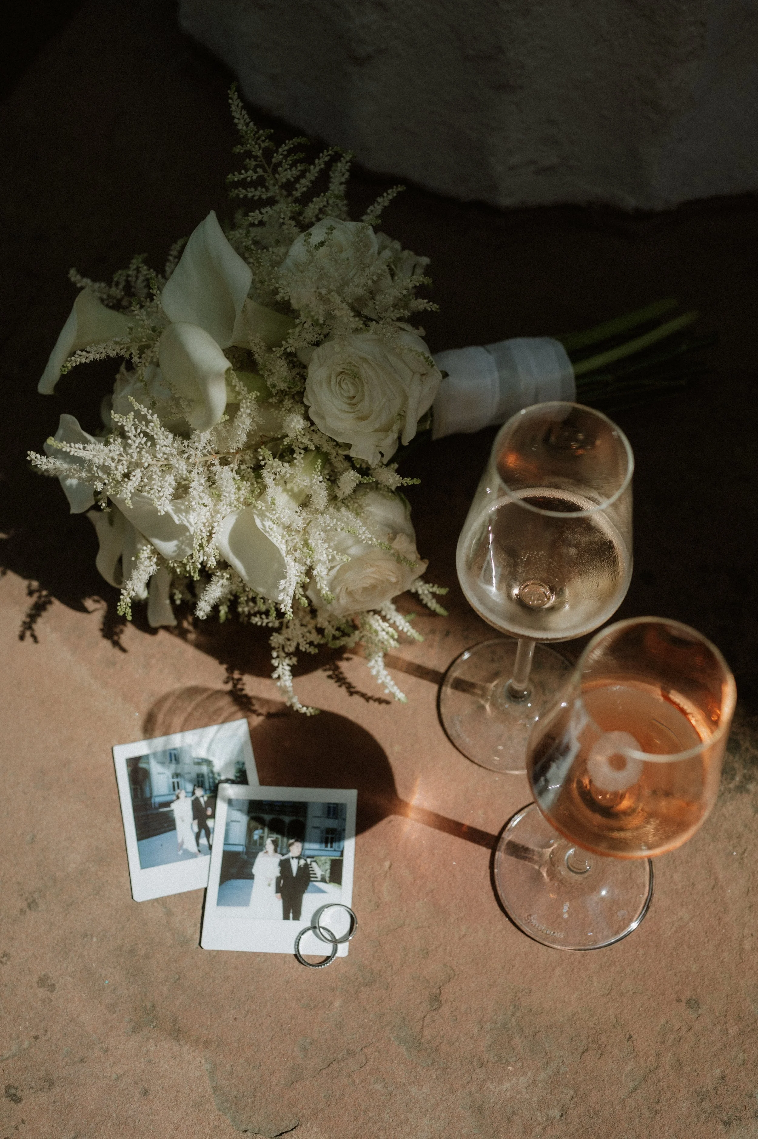 A wedding bouquet of white roses, calla lilies, and greenery, two glasses of rosé wine, and two wedding rings resting on a soft surface with two Polaroid photos placed nearby.