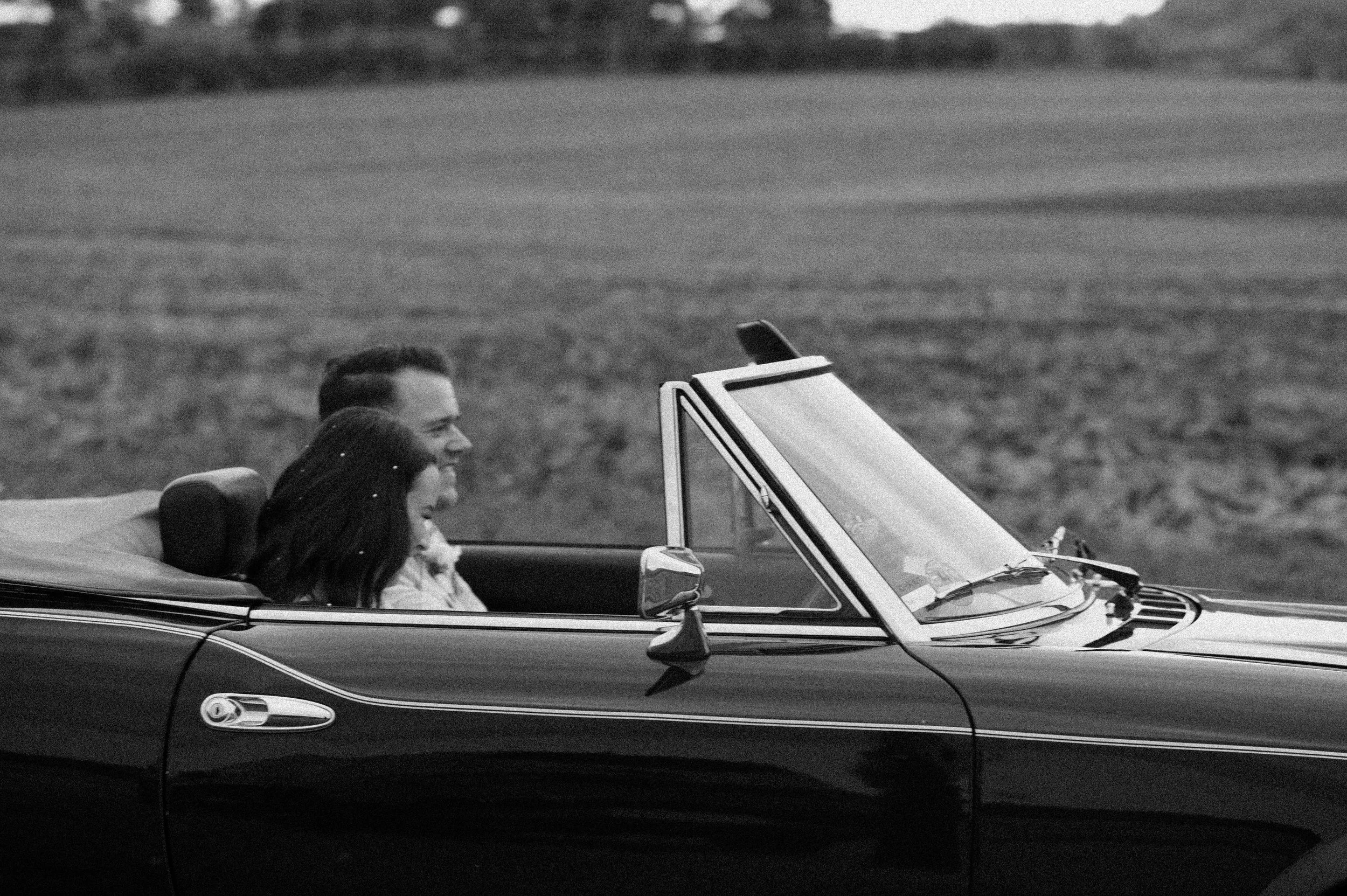 A black and white photo of a man and a young girl sitting in a vintage convertible car on a rural road, smiling and looking ahead.
