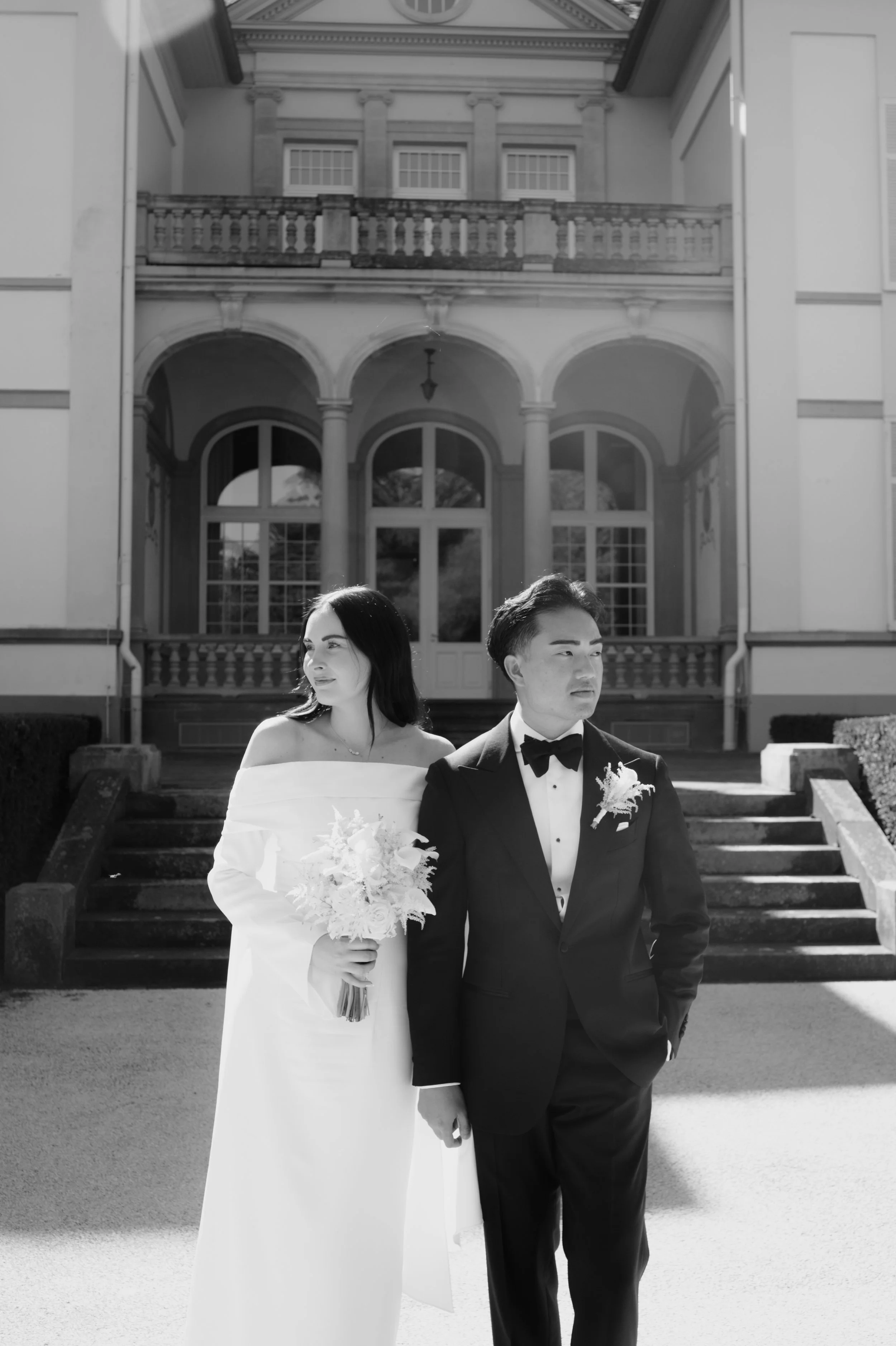 Black and white photo of a bride in an off-shoulder dress holding a bouquet, standing next to a groom in a tuxedo with a bow tie, in front of a large, ornate house with steps and columns.