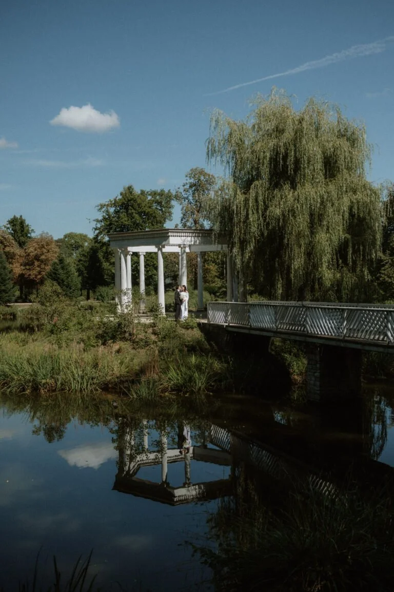 A couple in wedding attire standing under a white classical pavilion with columns, near a pond with a reflection, surrounded by trees and greenery on a sunny day.