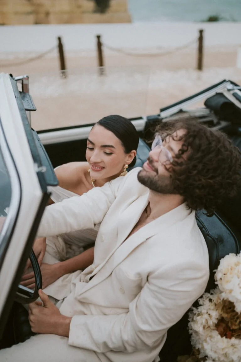 A man and woman sitting in a convertible car, smiling and enjoying each other's company outside during daytime, with a stone wall and chain barrier in the background.