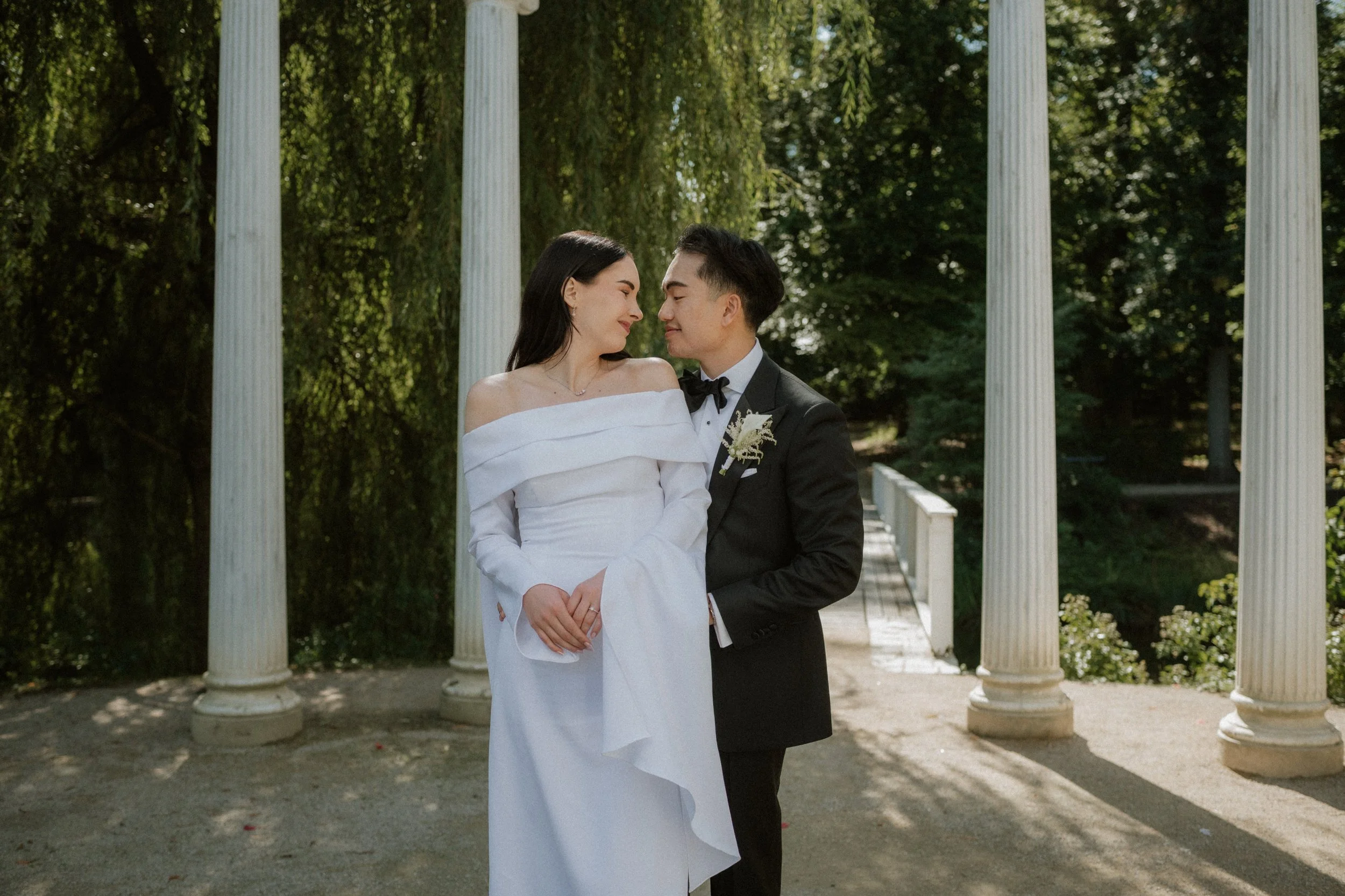 A bride and groom standing close together outdoors, smiling at each other beneath a white gazebo with pillars, surrounded by green trees.