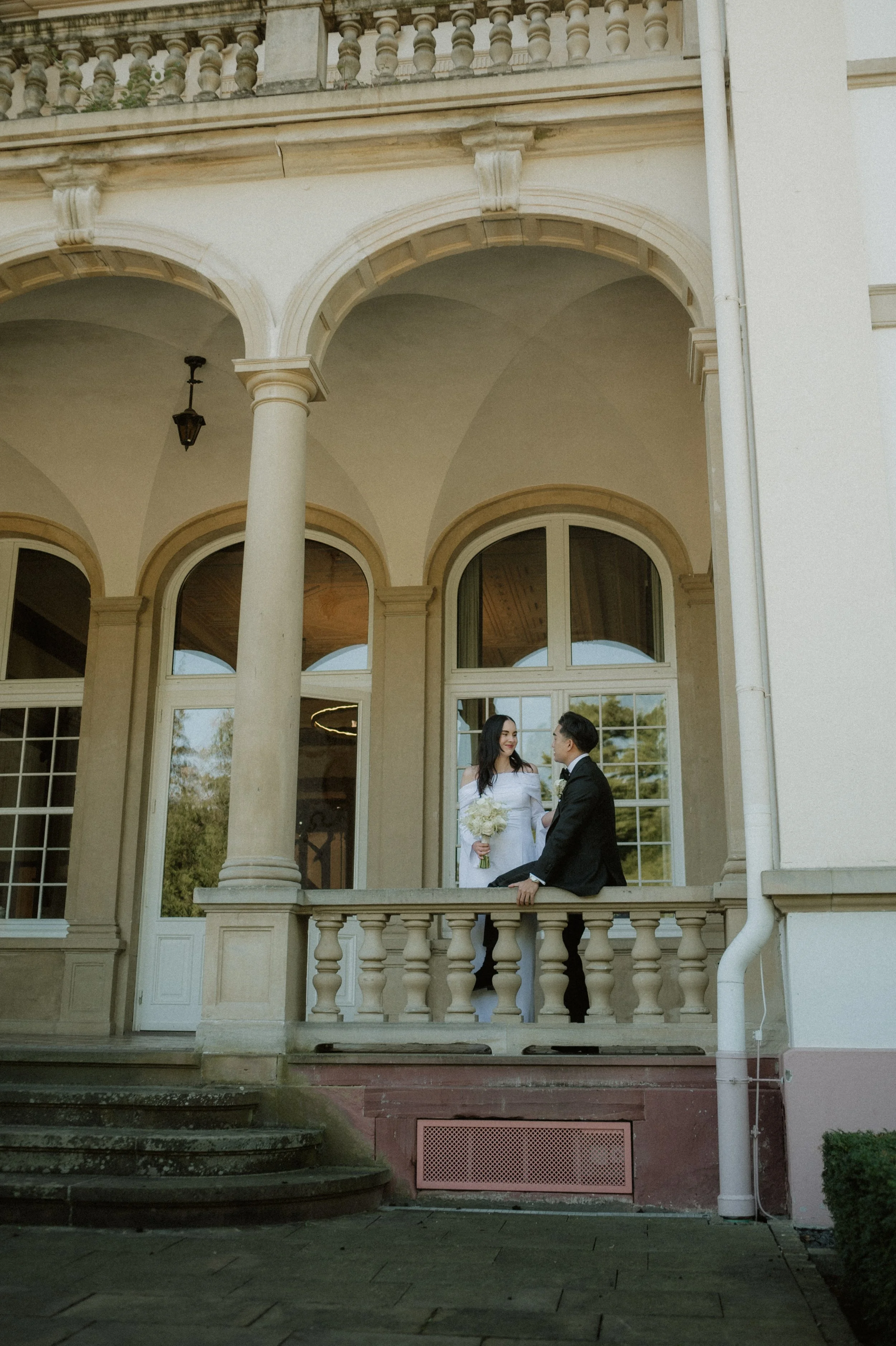 A bride and groom standing on a porch of a large, elegant house with arched windows and columns, holding a bouquet of flowers, sharing a moment.