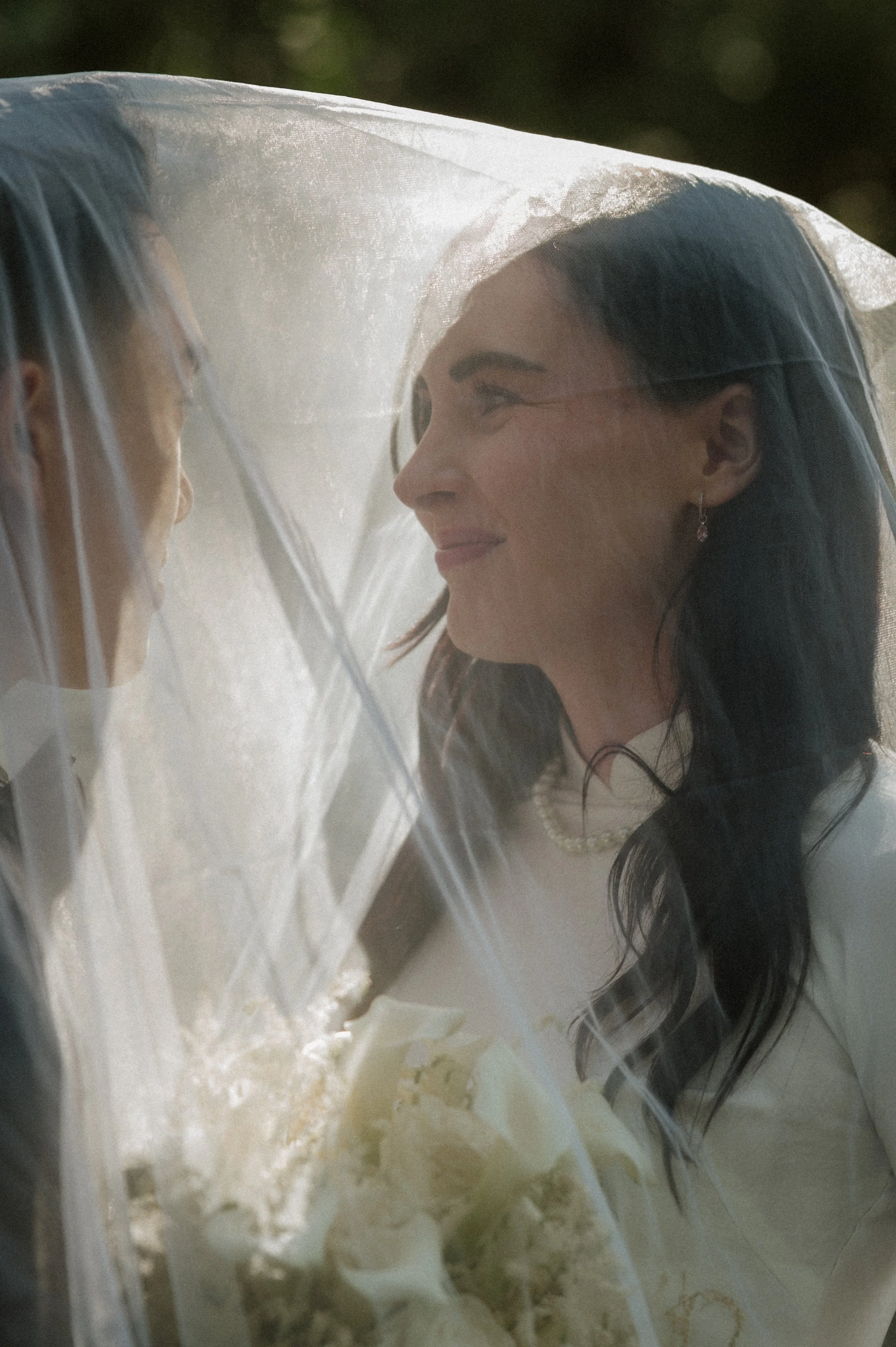 A bride and groom face each other under a sheer wedding veil, with the bride smiling lovingly.