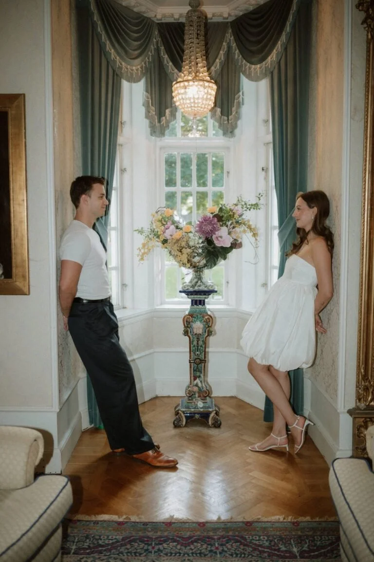 A man and woman stand facing each other in a decorated room with draped curtains and a chandelier. The woman wears a white dress and heels, and the man wears a white shirt and dark pants. They stand on either side of a tall floral arrangement on an ornate pedestal.