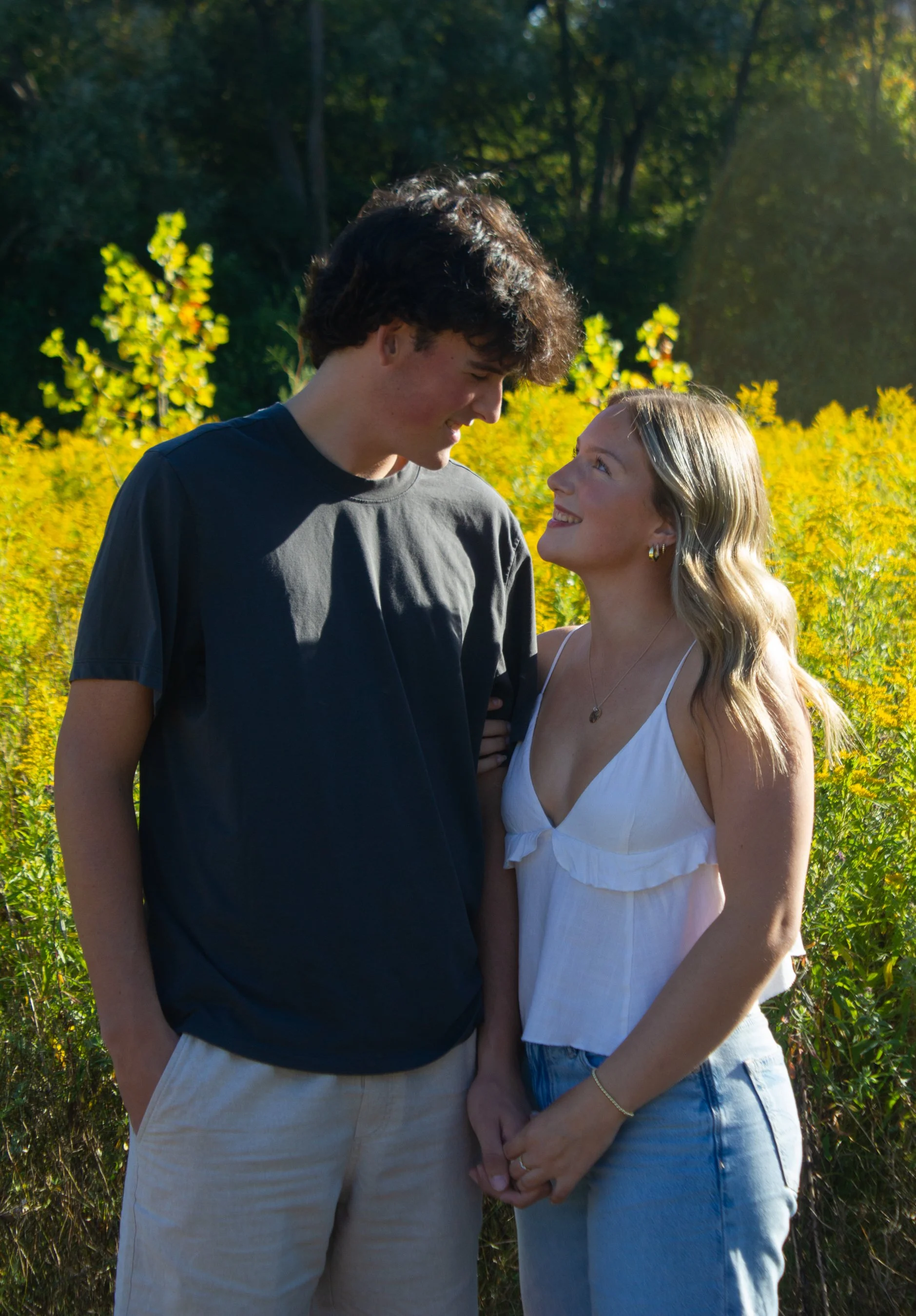A young man and woman holding hands and smiling at each other outdoors in a field of yellow flowers with green trees in the background.