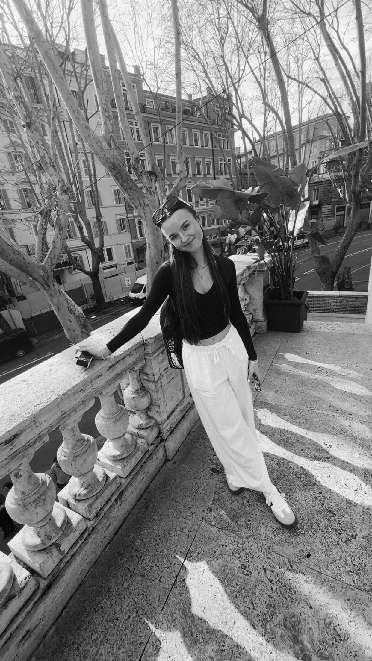 A young woman with long hair standing outdoors on a balcony, leaning on a stone railing, with urban buildings and leafless trees in the background. She is wearing a black long-sleeve top and white pants, and holding a phone in her right hand.