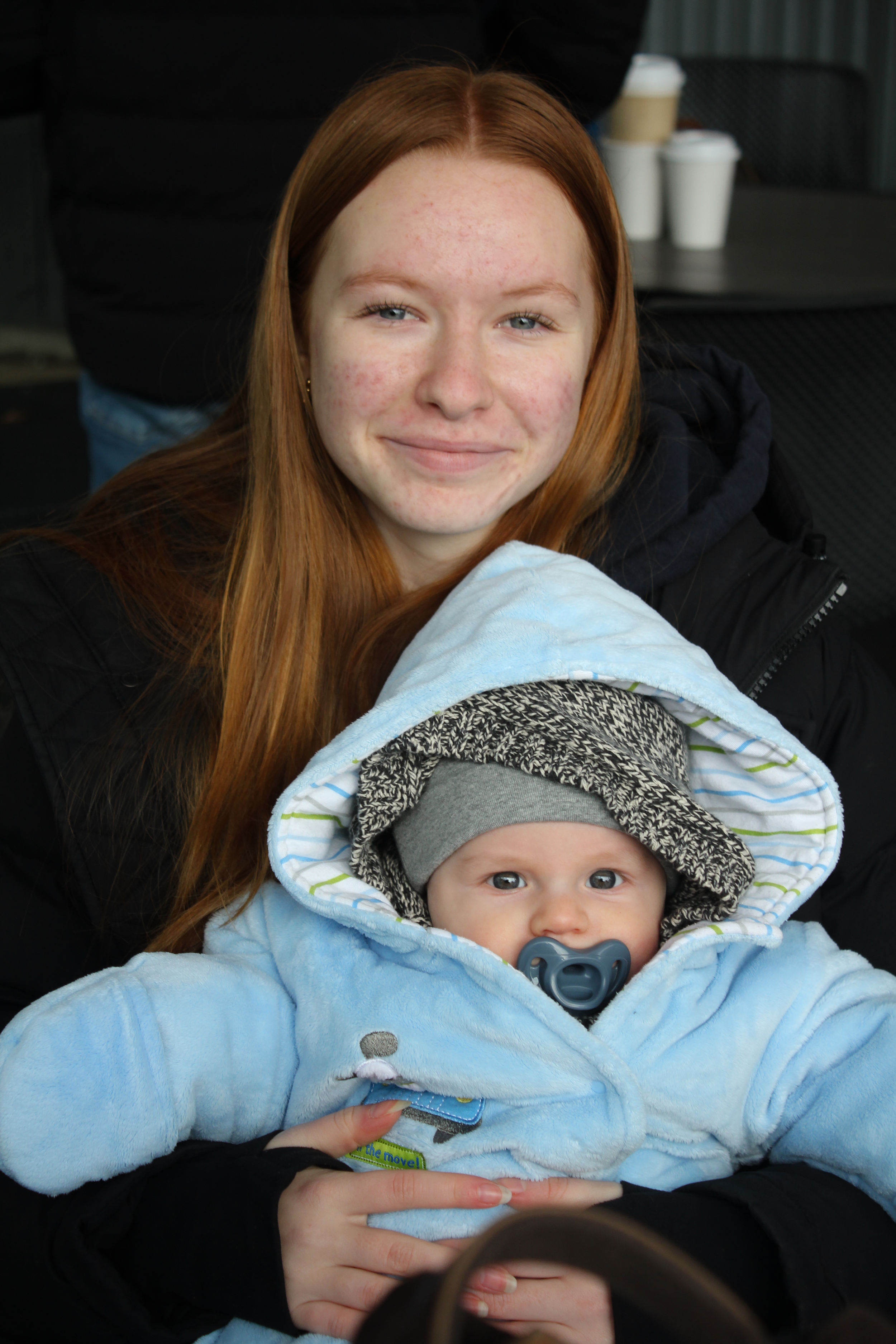 A young woman with long red hair and a baby with pacifier in a blue hoodie, sitting together, smiling at the camera.