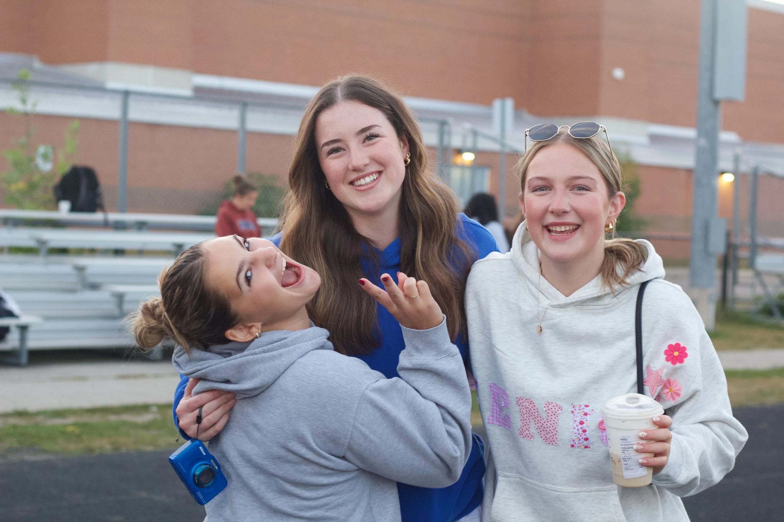 Three young girls happily posing outdoors at a sports field or stadium, with metal bleachers and a brick building in the background. One girl is wearing glasses on her head and holding a drink, another has a camera hanging around her neck, and they are all smiling and having fun.