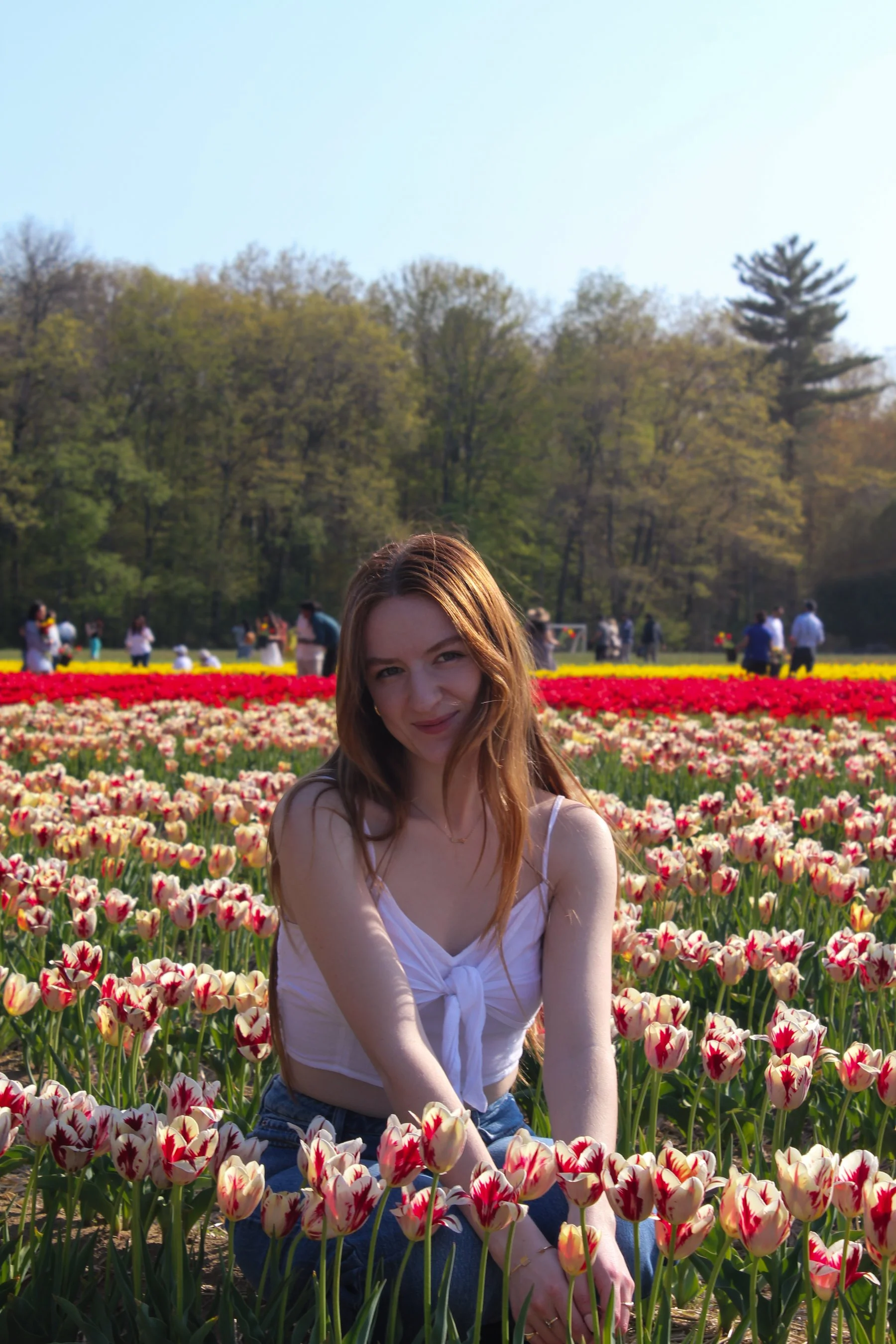 Young woman with red hair in a white top sitting among colorful tulips in a flower garden on a sunny day.