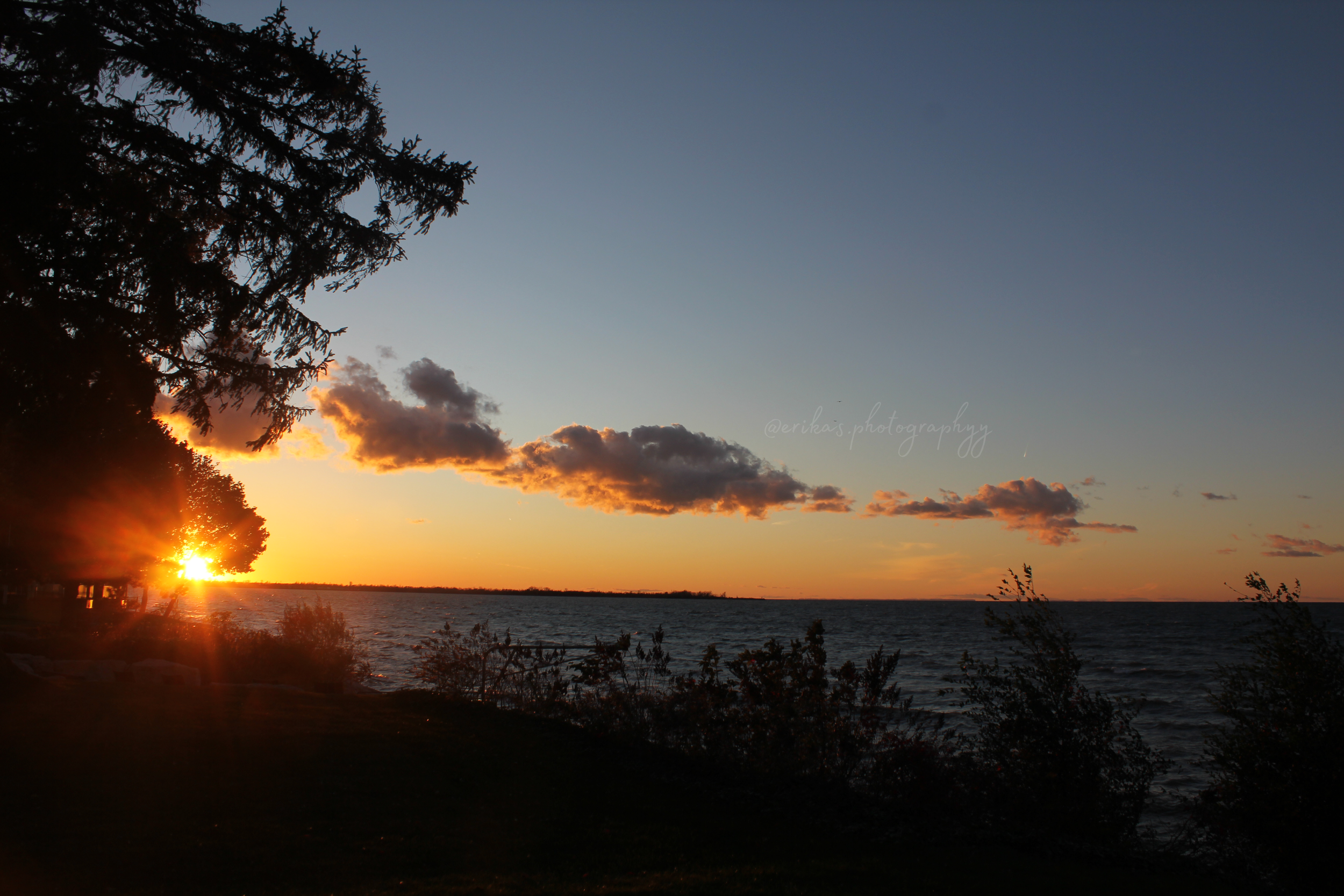 Sunset over a lake seen through trees with a partly cloudy sky.