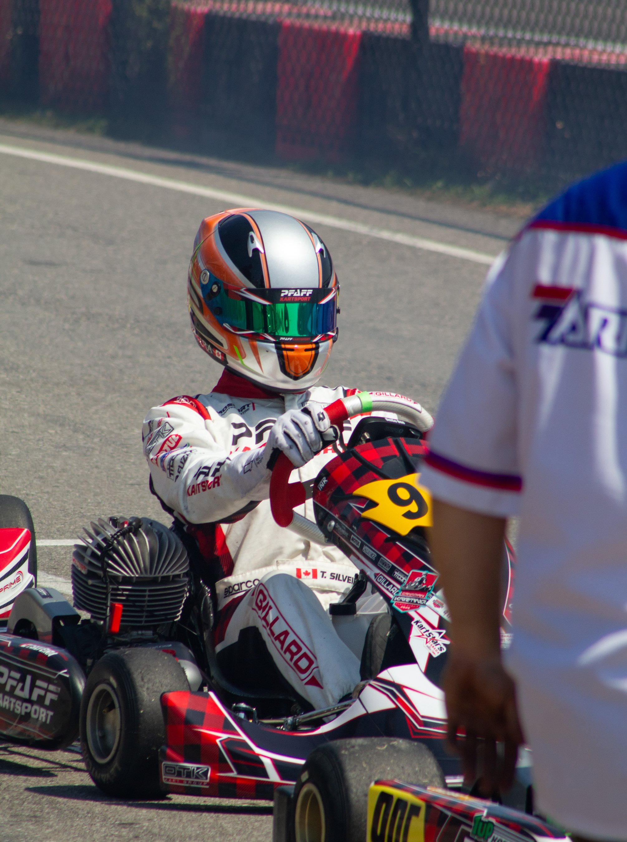 A go-kart racer in a racing suit and helmet sits in a motorized kart on a racetrack. The person is holding the steering wheel and appears to be getting instructions from another individual standing nearby.