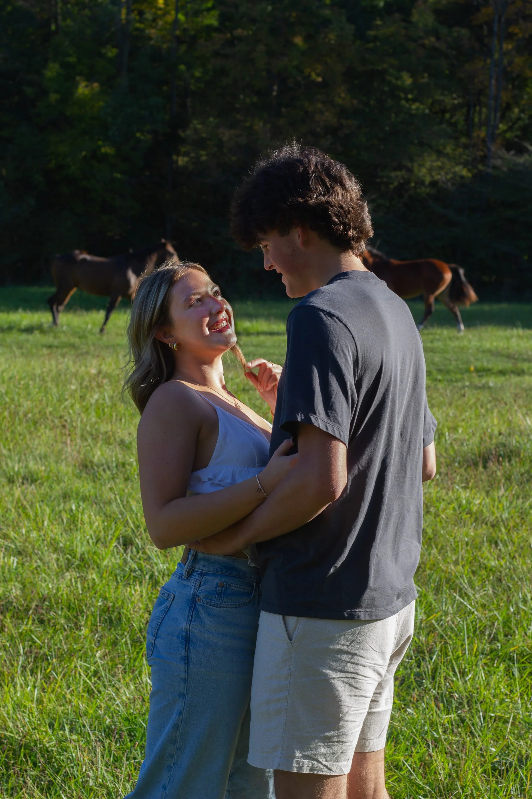 A young woman and man standing close together in a grassy field, smiling and gazing into each other's eyes, with two horses grazing in the background and trees beyond.