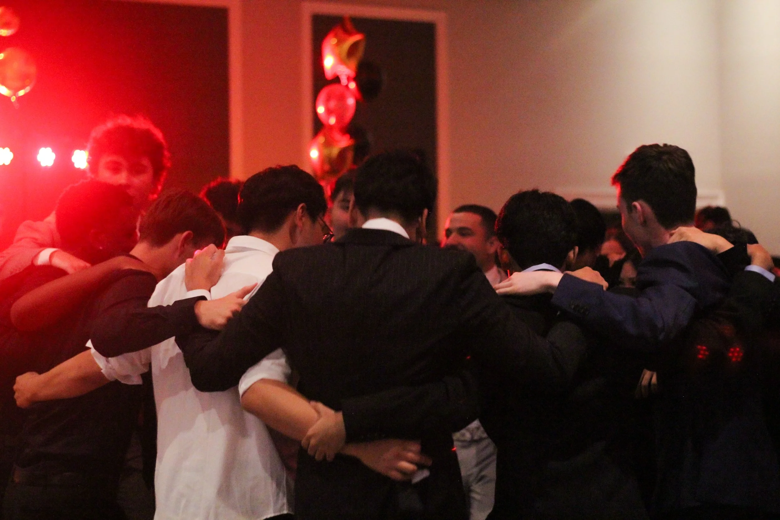 Group of young men in suits and formal attire huddled together in a circle, possibly at a party or celebration, with red lighting and balloons in the background.